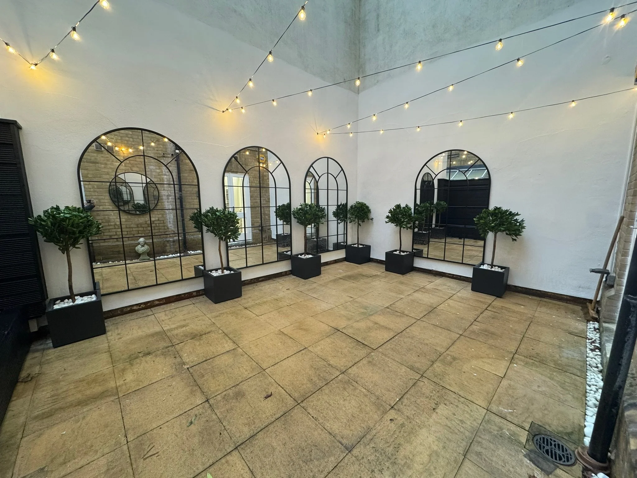 A patio space with string lights overhead, three potted trees aligned along the wall, and four large mirrors with black metal frames leaning against the white wall.