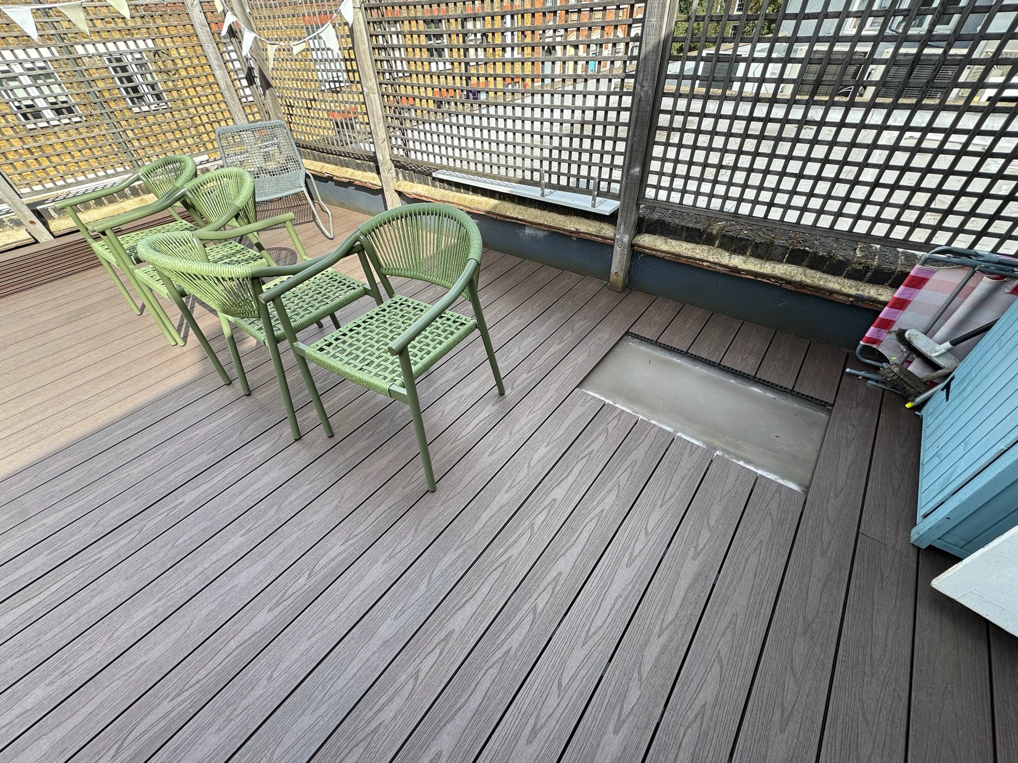 A rooftop balcony with wooden flooring, four green chairs, a small gray table, and a storage cabinet. The background has a lattice fence and cityscape view.