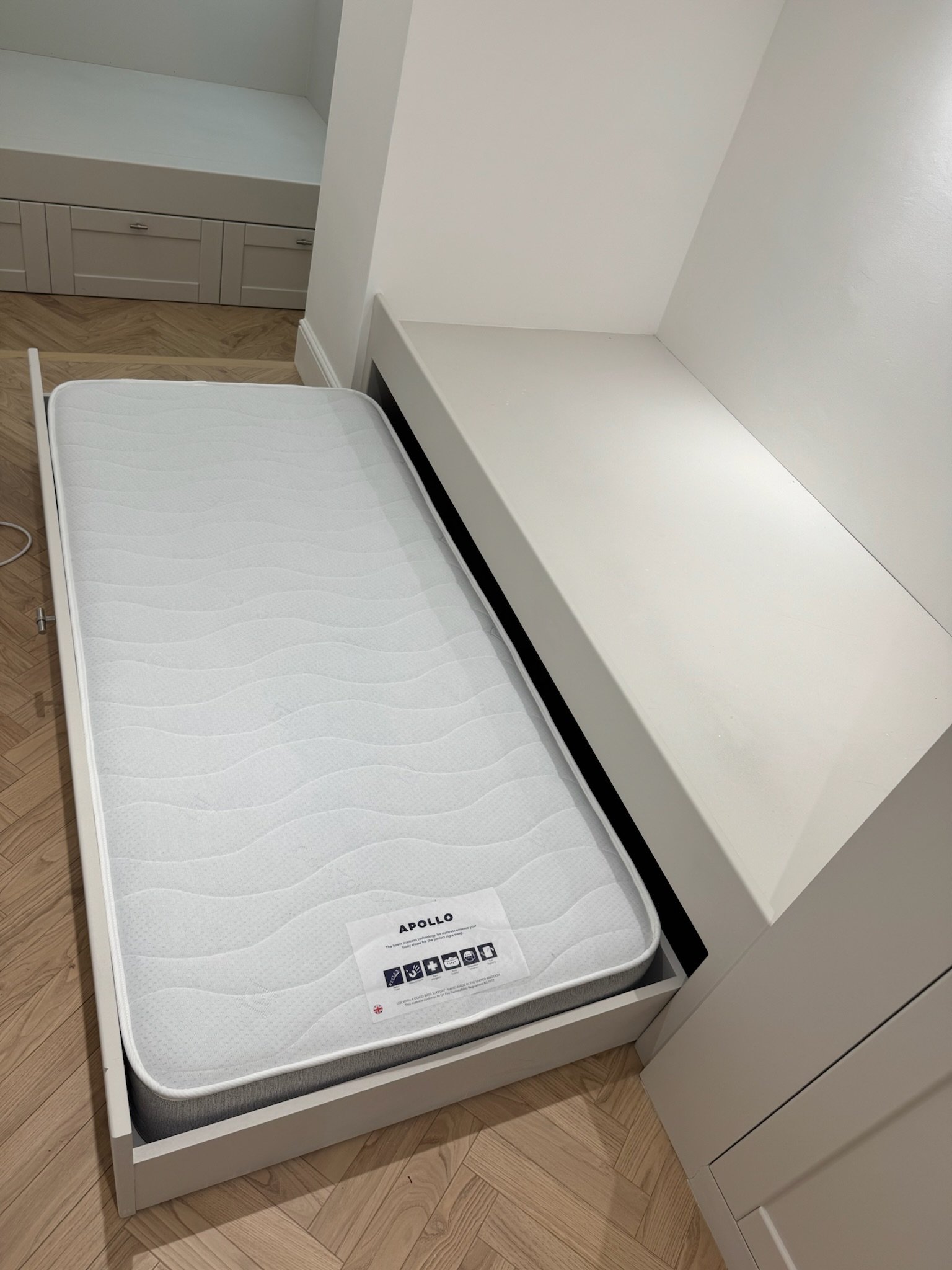 A white mattress in a partially pulled out bed frame next to a white built-in bench in a room with light wood flooring and a white wall.