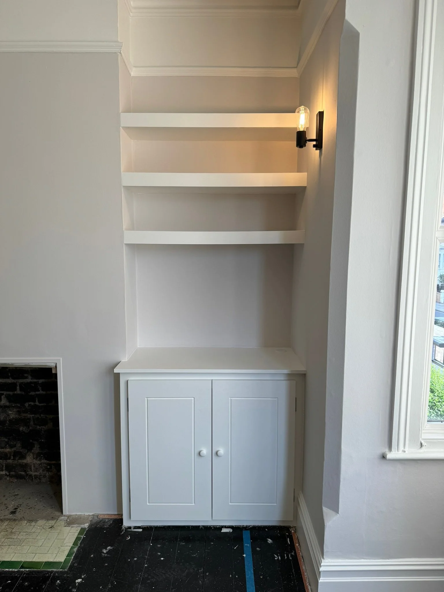 White built-in shelving unit with three shelves above a cabinet, located next to a window and a wall sconce with an exposed bulb, within a room under renovation.
