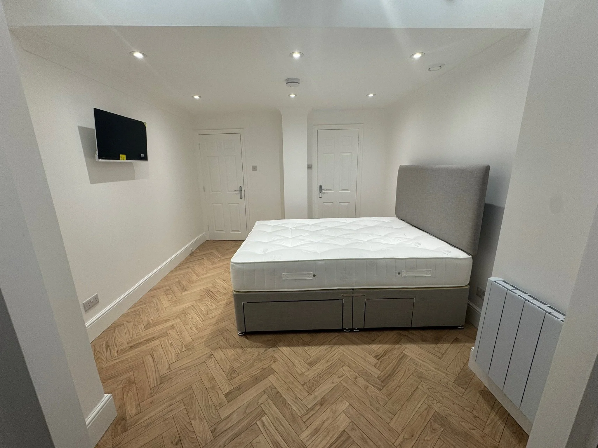 A minimalist bedroom with a bed that has a gray upholstered headboard and a white mattress, wooden flooring, a wall-mounted flat-screen TV, and two white doors.