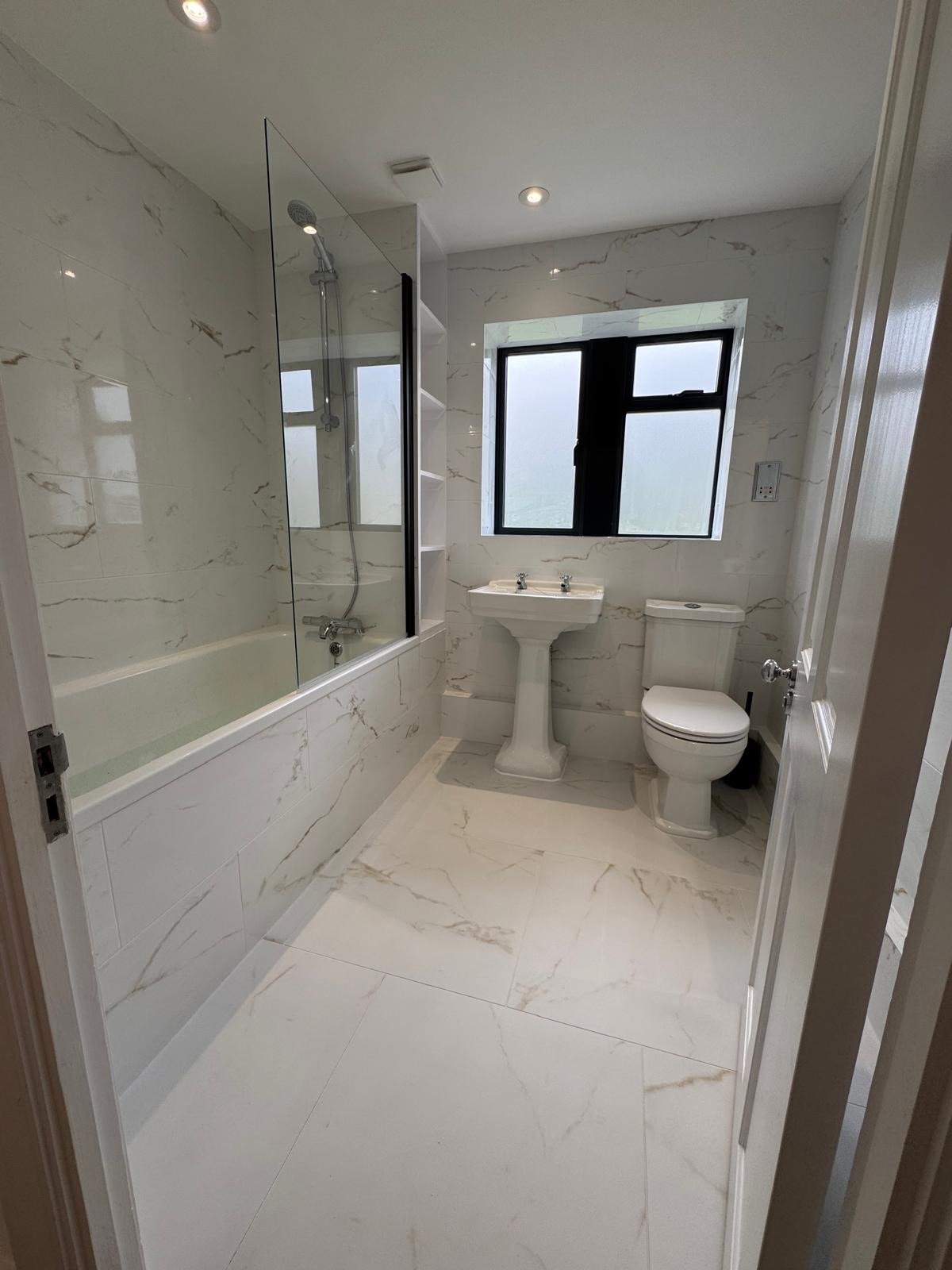 Bathroom with white marble walls and floor, bathtub with shower and glass partition, pedestal sink, toilet, black window, and ceiling lights.