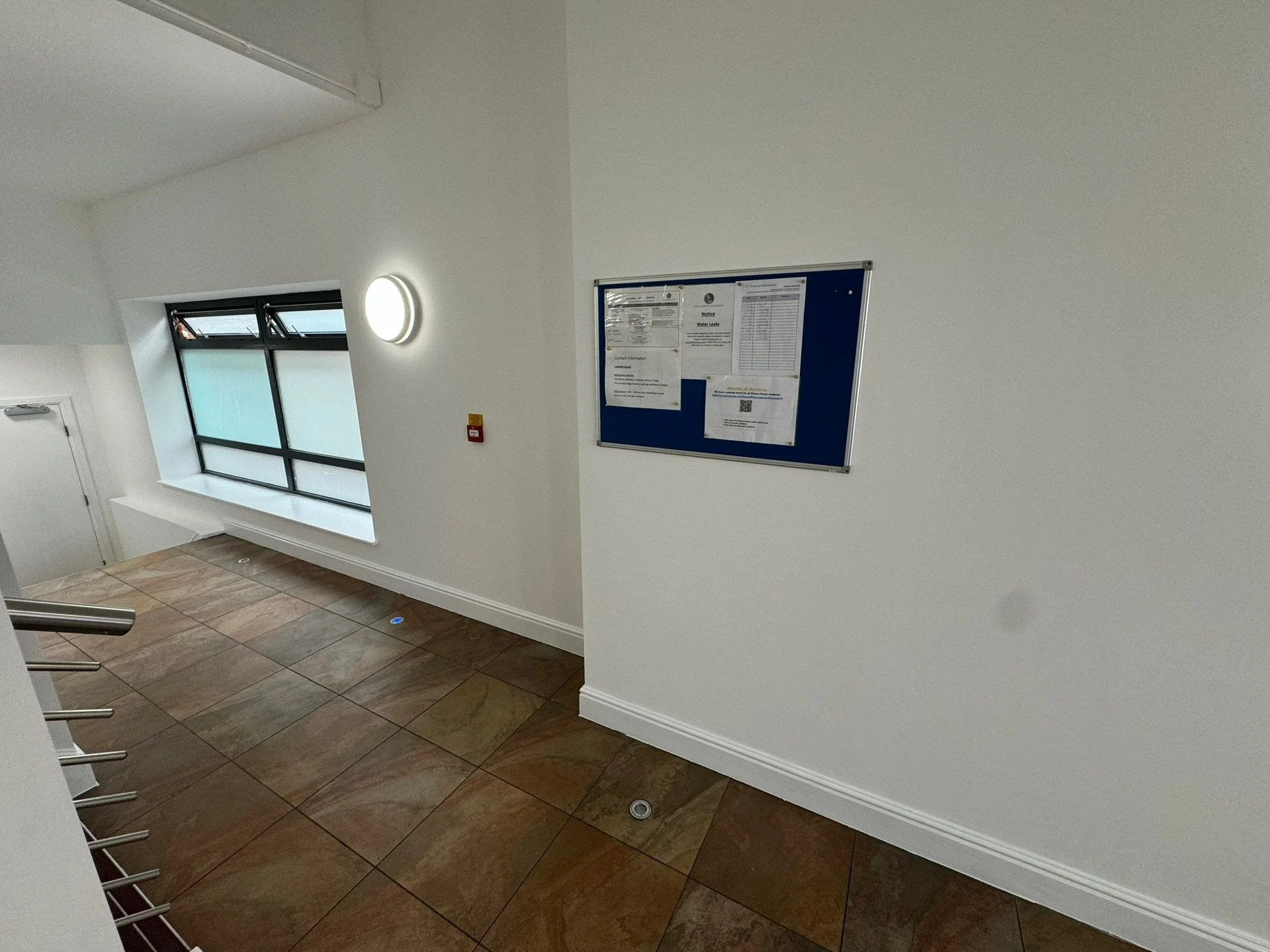 Interior of a stairwell landing with a large window, wall-mounted light, blue notice board, red fire alarm, white walls, and tiled floor.