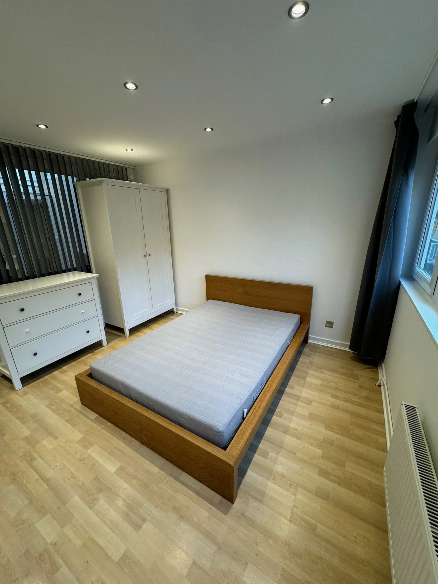 Minimalist bedroom with a wooden bed frame, a white dresser, a white wardrobe, a window with black curtains, and recessed ceiling lights.