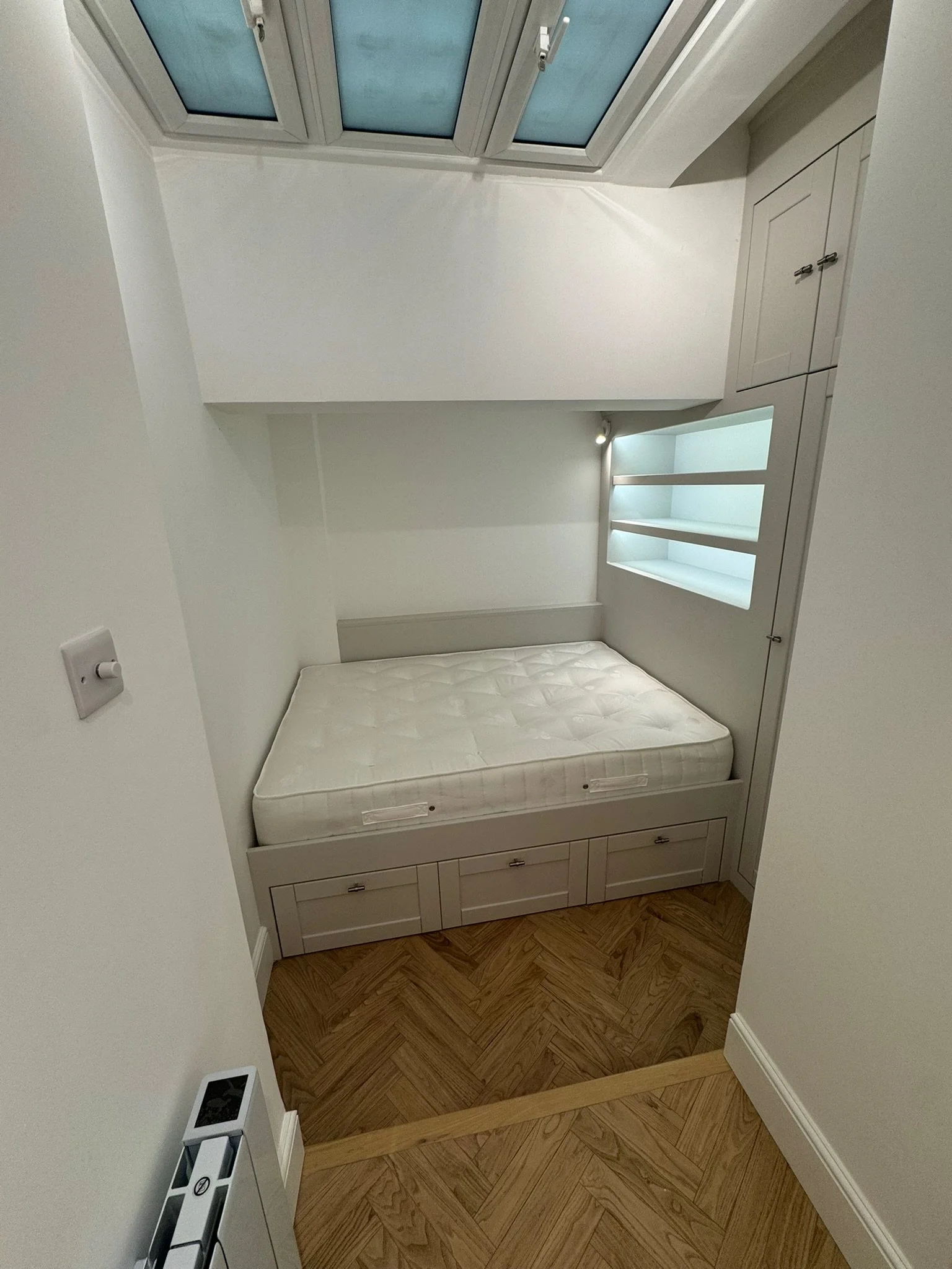 Small bedroom with white walls, a windowed ceiling, built-in white furniture, a bed with a mattress, and wooden parquet flooring.