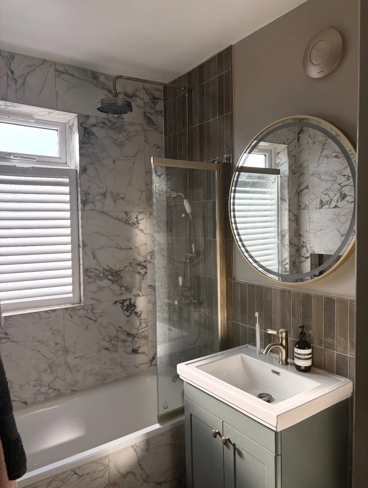 Bathroom with marble walls, a bathtub with a glass shower barrier, a window with white blinds, a round mirror, a gray sink cabinet, a faucet, and soap dispenser.