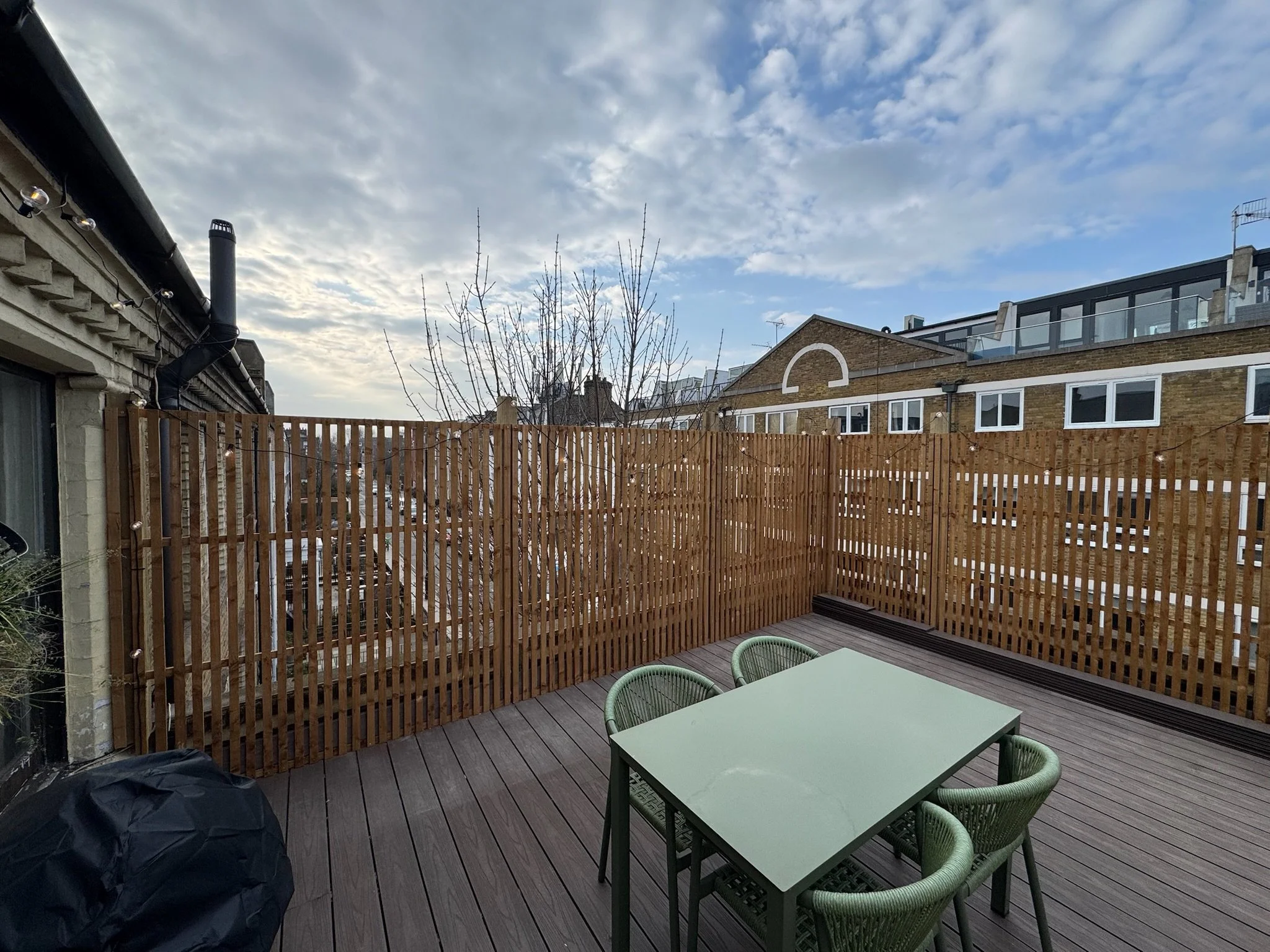 A small urban rooftop patio with wooden decking, a green table, four green chairs, a black cover on an object, a wooden fence, leafless tree, neighboring buildings with windows, and a partly cloudy sky