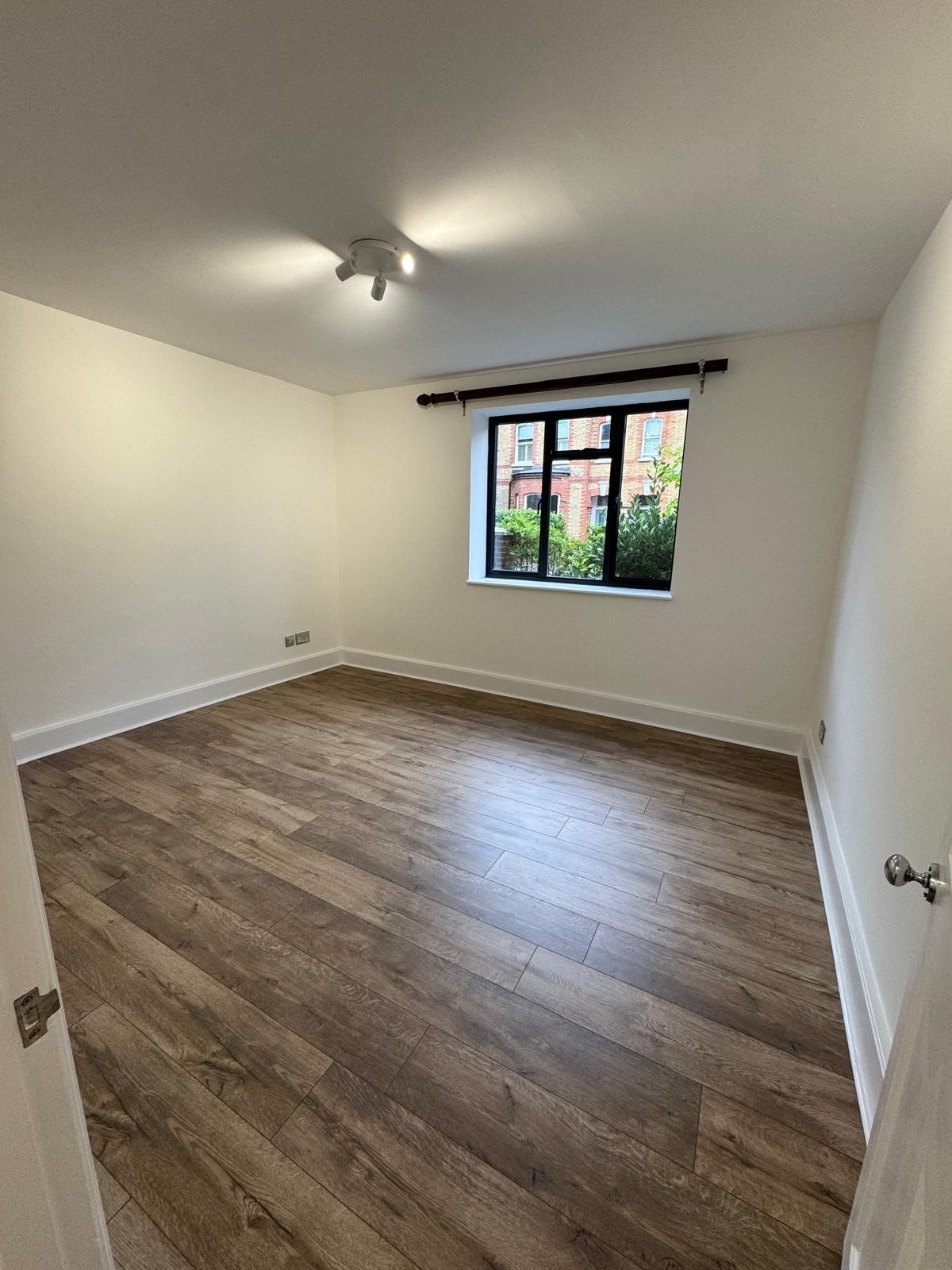 Empty room with white walls, wooden flooring, a single window with a black frame, and a ceiling light fixture.