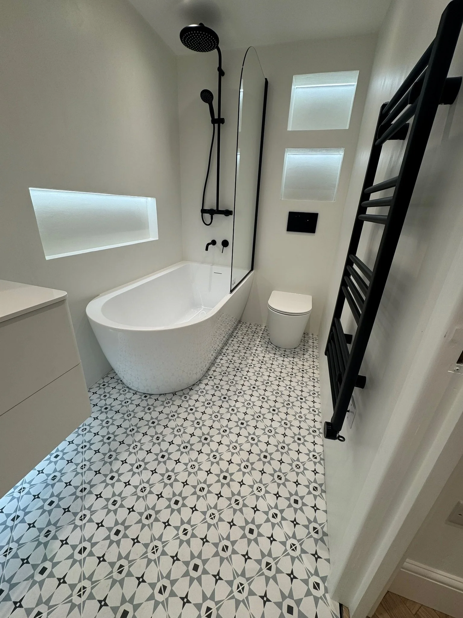 Modern bathroom featuring a freestanding bathtub with a glass shower screen, a black shower head and fixtures, patterned black and white tile floor, a wall-mounted toilet, black towel heating rack, and minimal white walls with two narrow high windows