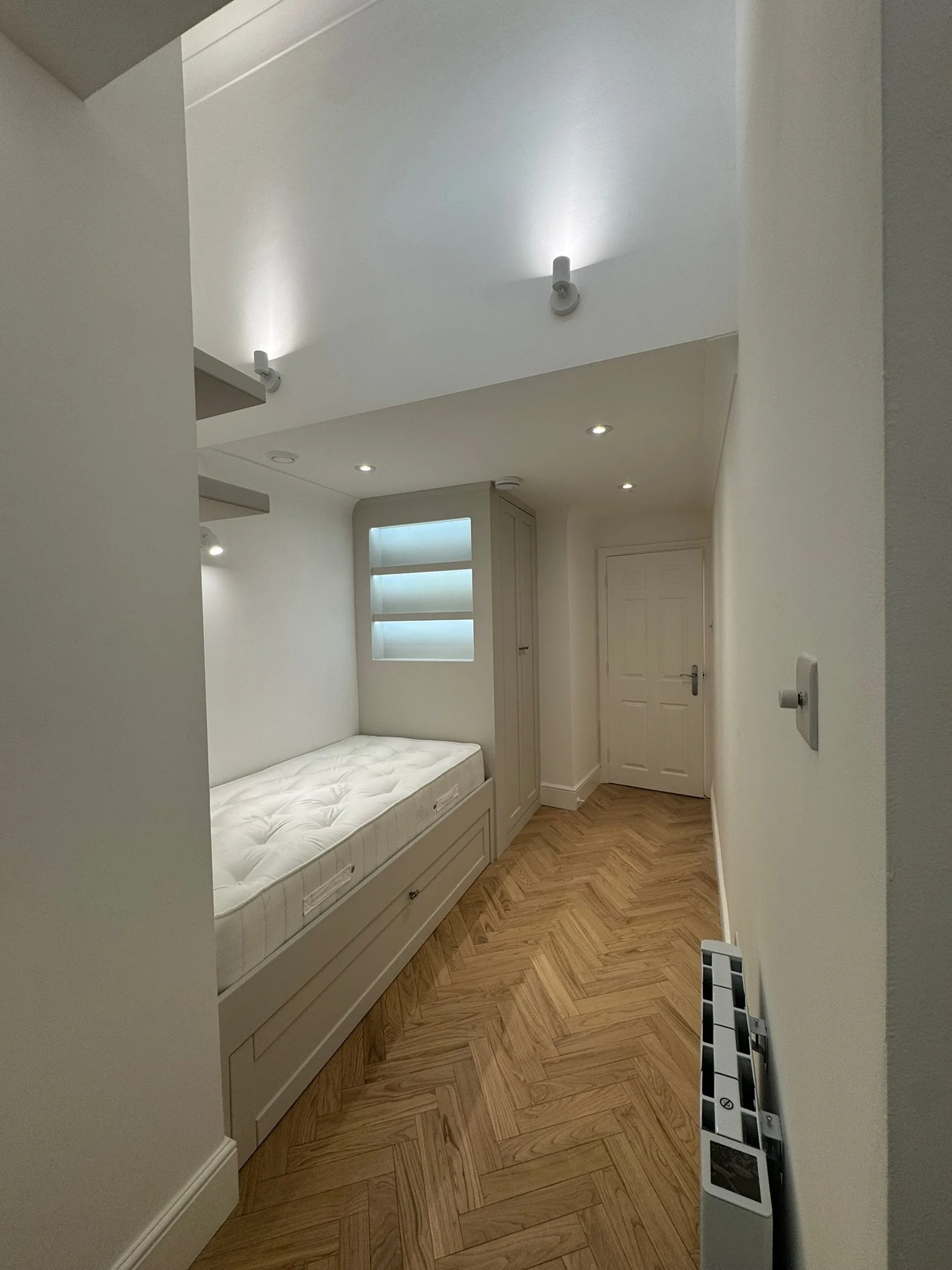 A small, modern bedroom with a white bed and built-in wardrobe, herringbone wood flooring, and bright ceiling lights.