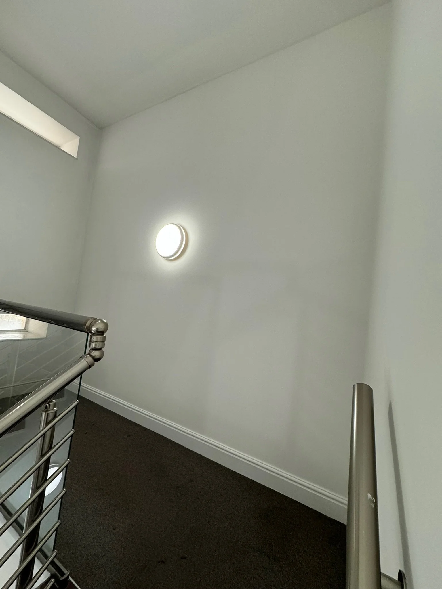Interior staircase landing with metal handrail, white walls, carpeted floor, and a round wall-mounted light fixture.
