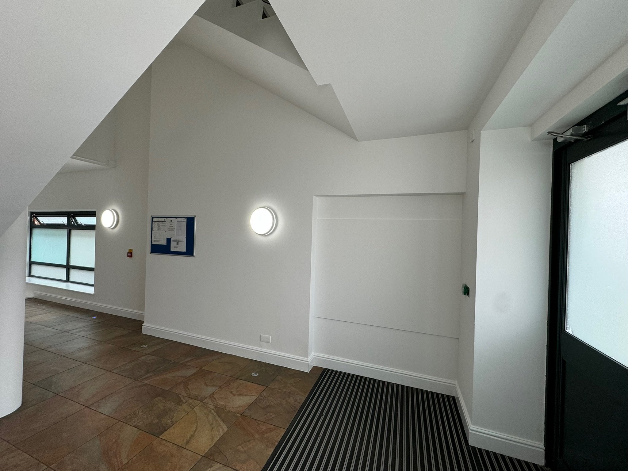 Interior of a modern building with white walls, a sloped ceiling, a window with black frames, circular wall lights, a notice board, and a black door with frosted glass.