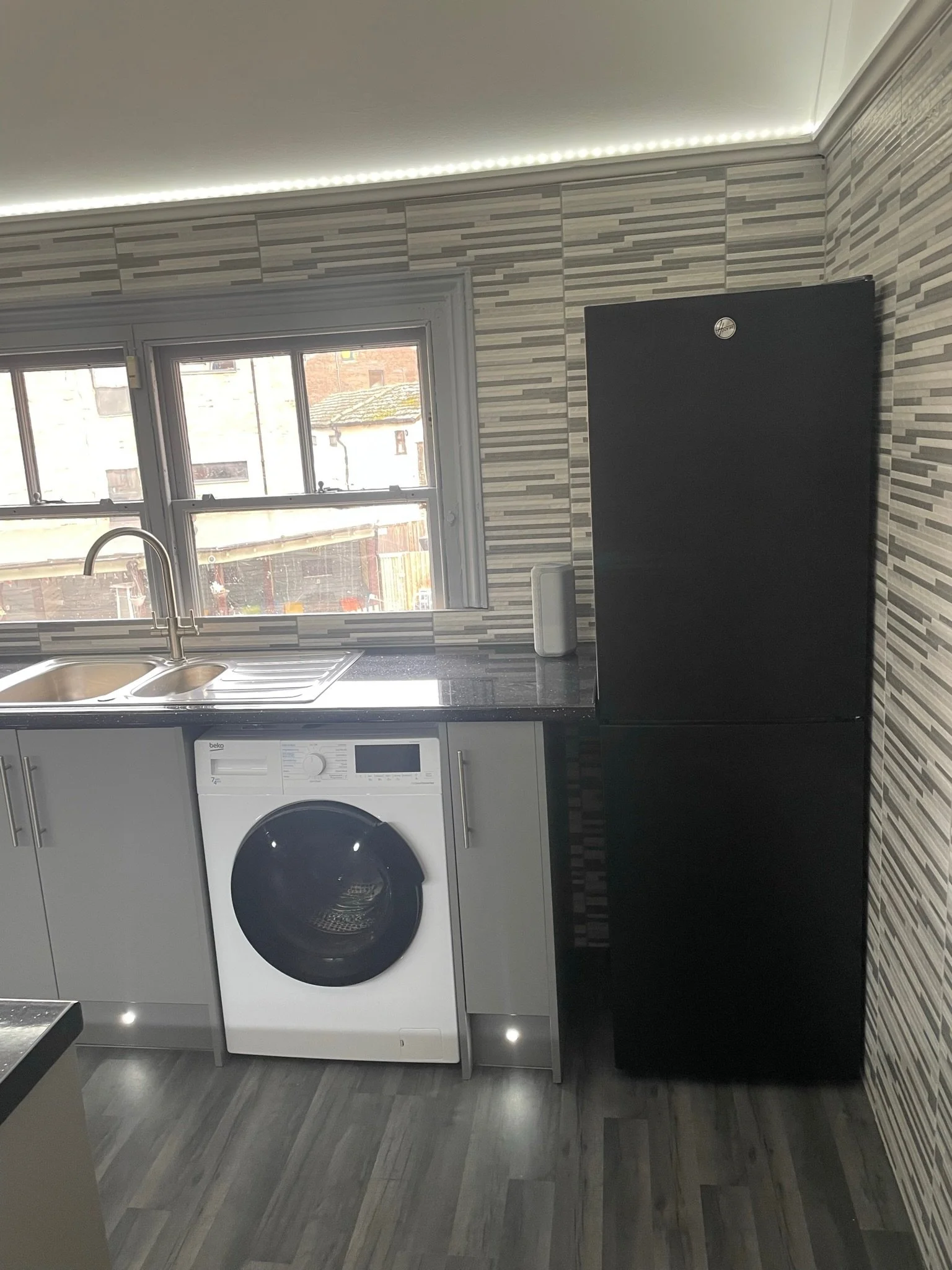 A kitchen with a black refrigerator, a washing machine, a window, a sink, and cabinets with a striped tile backsplash.
