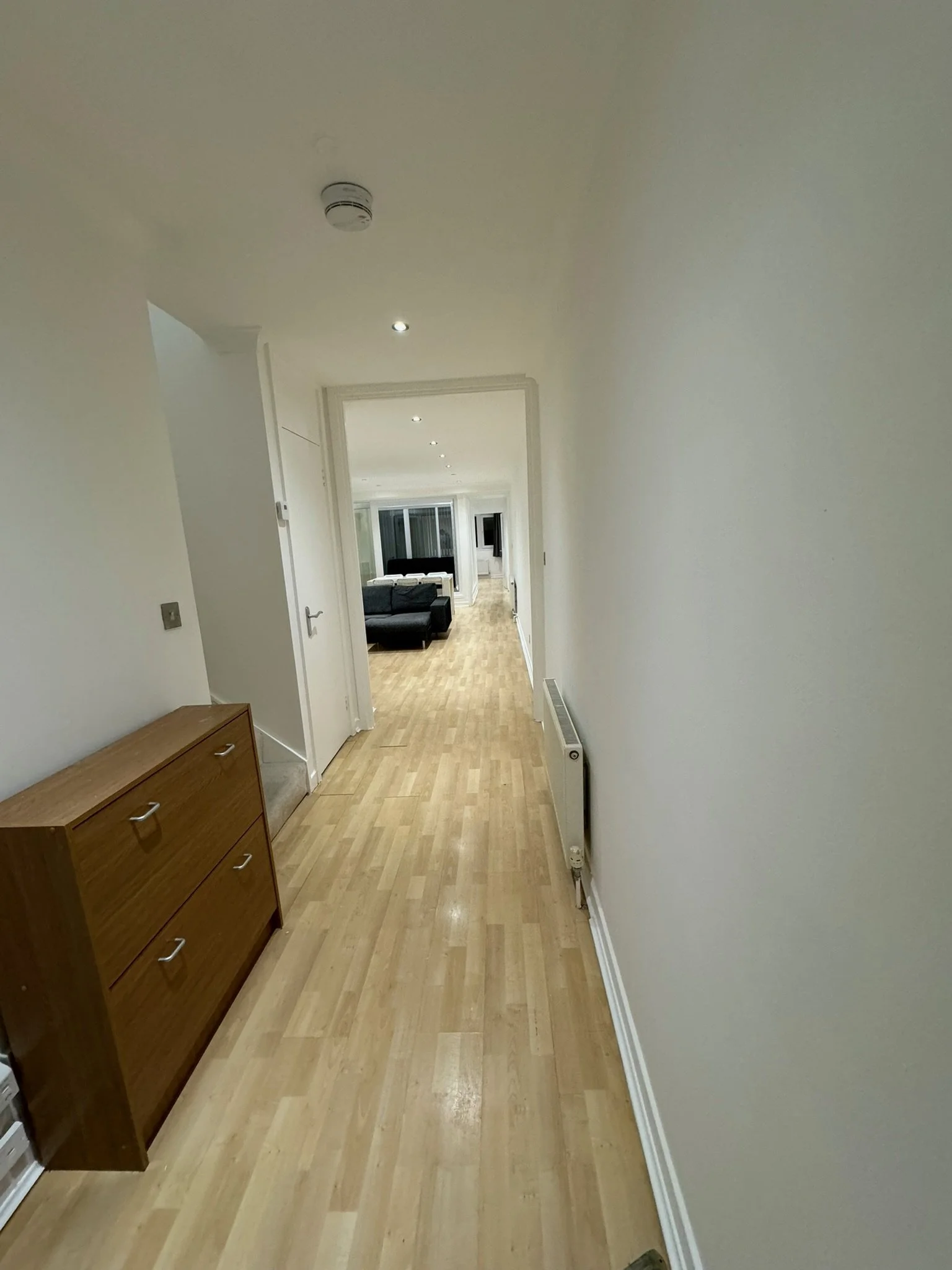 Empty hallway with wooden flooring, white walls, a small wooden cabinet on the left, and a radiator on the right, leading to a living room with a sofa and large windows.