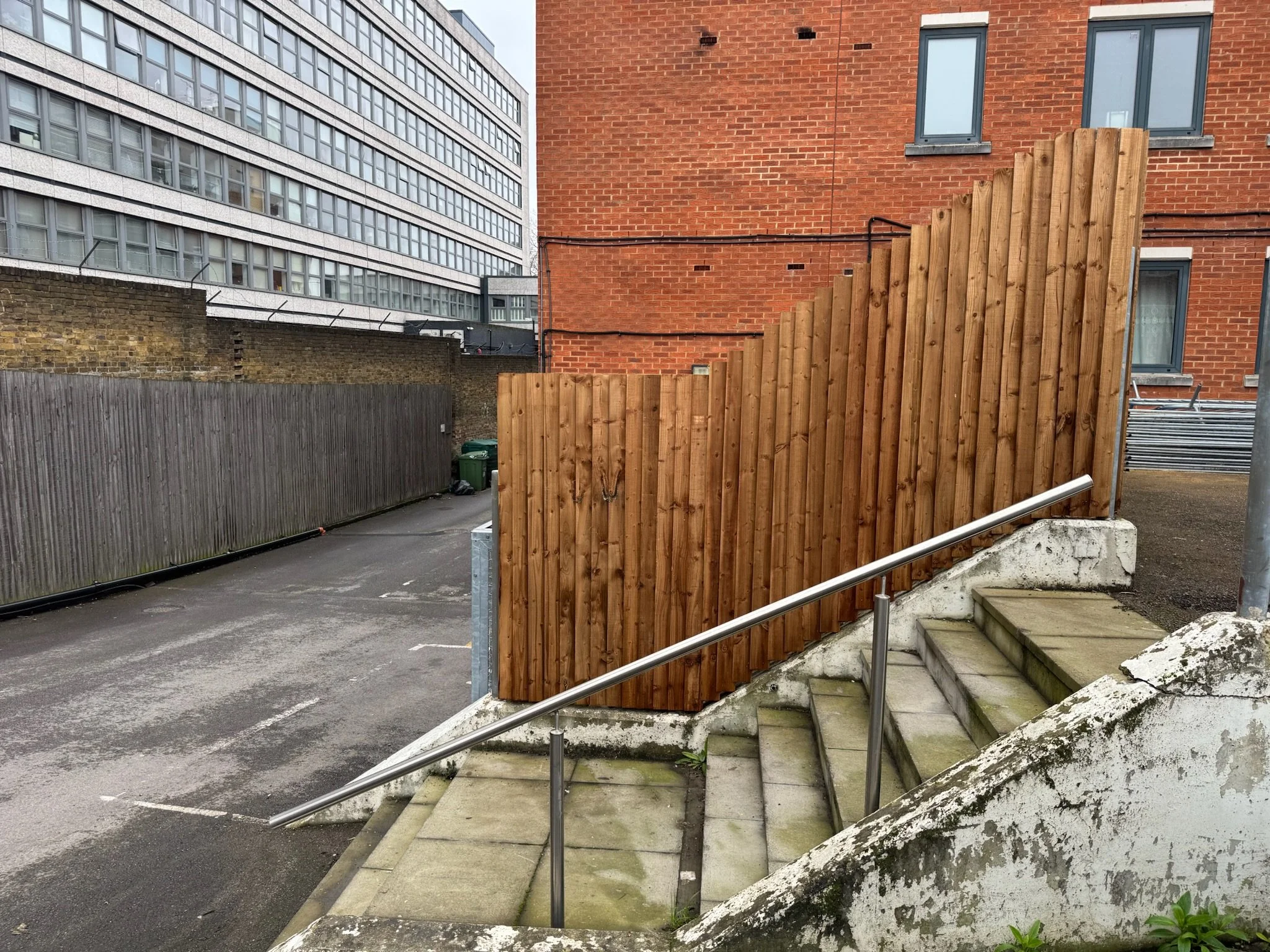 Wooden fence alongside a concrete staircase with a metal handrail, located in an urban parking area near brick and glass buildings.