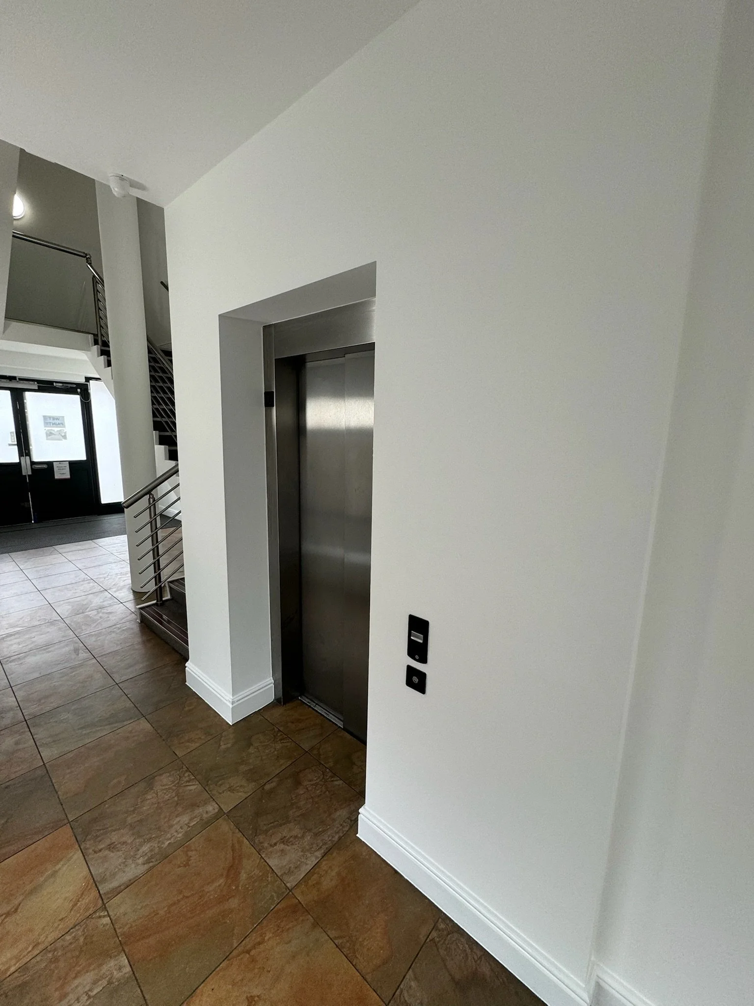 Interior view of an elevator with closed stainless steel doors, white walls, and tiled floor.
