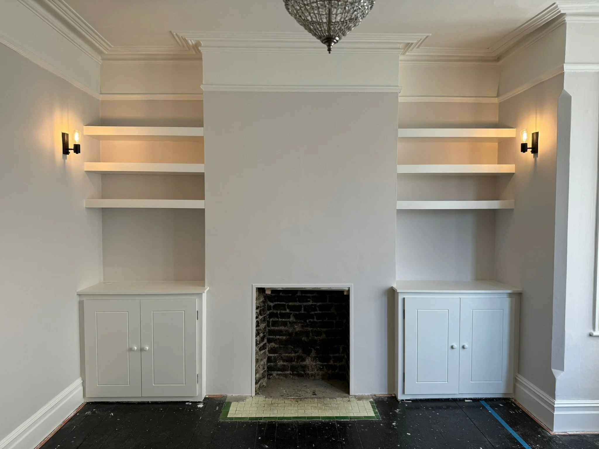 Interior of a room with built-in white cabinets and open shelves on either side of a central fireplace space, with wall-mounted lights, crown molding, and a decorative ceiling light fixture.