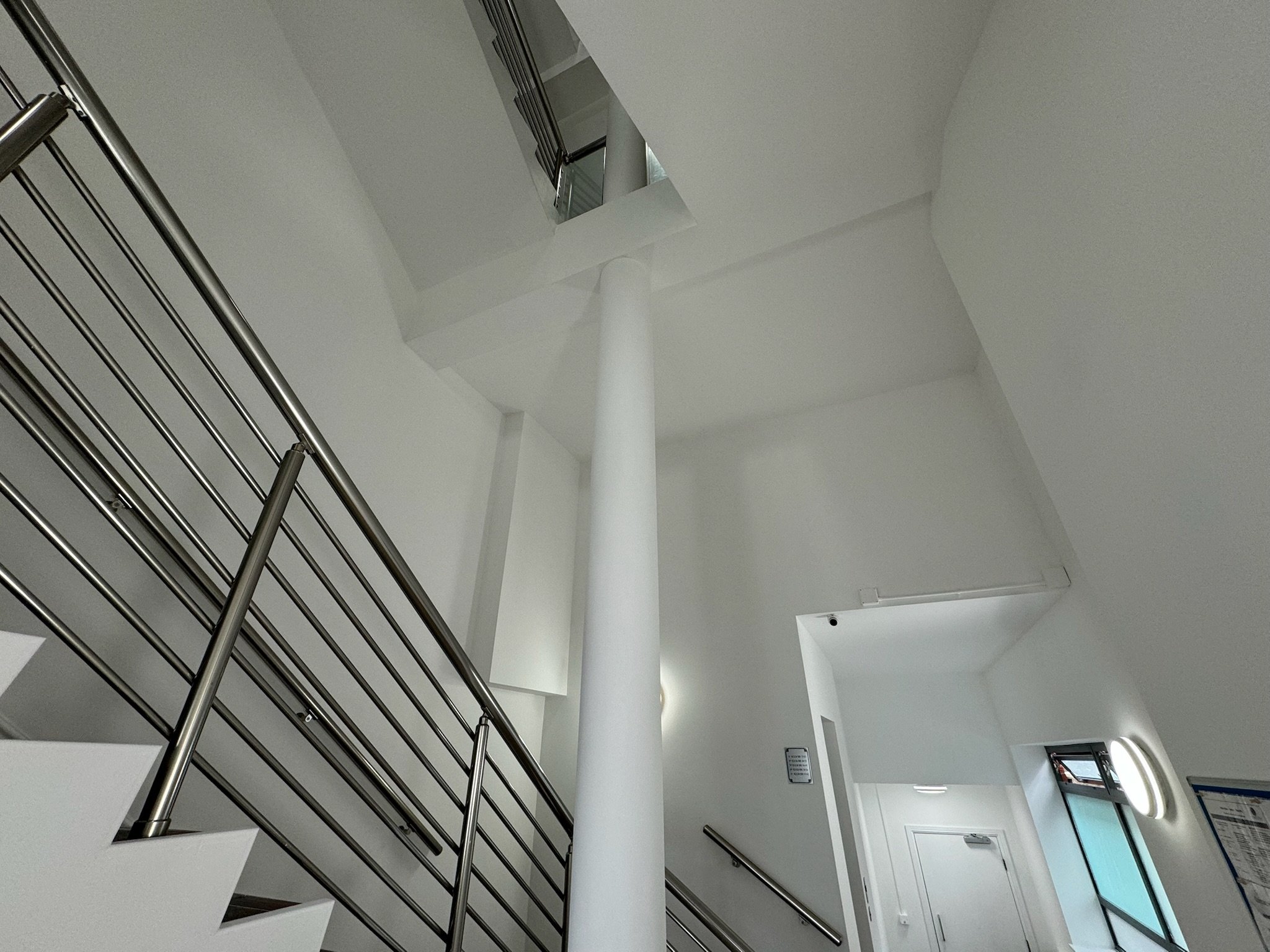 Interior view of a modern building with white walls, a staircase with metallic railings, a central white pillar, and a window allowing natural light.