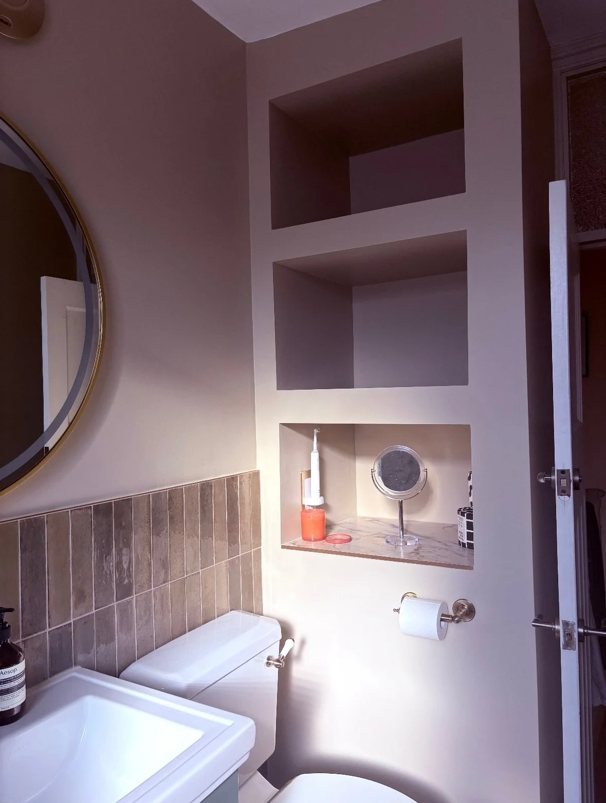Part of a bathroom showing a mirror, a toilet, a small sink, and built-in wall shelves with various toiletries and items.