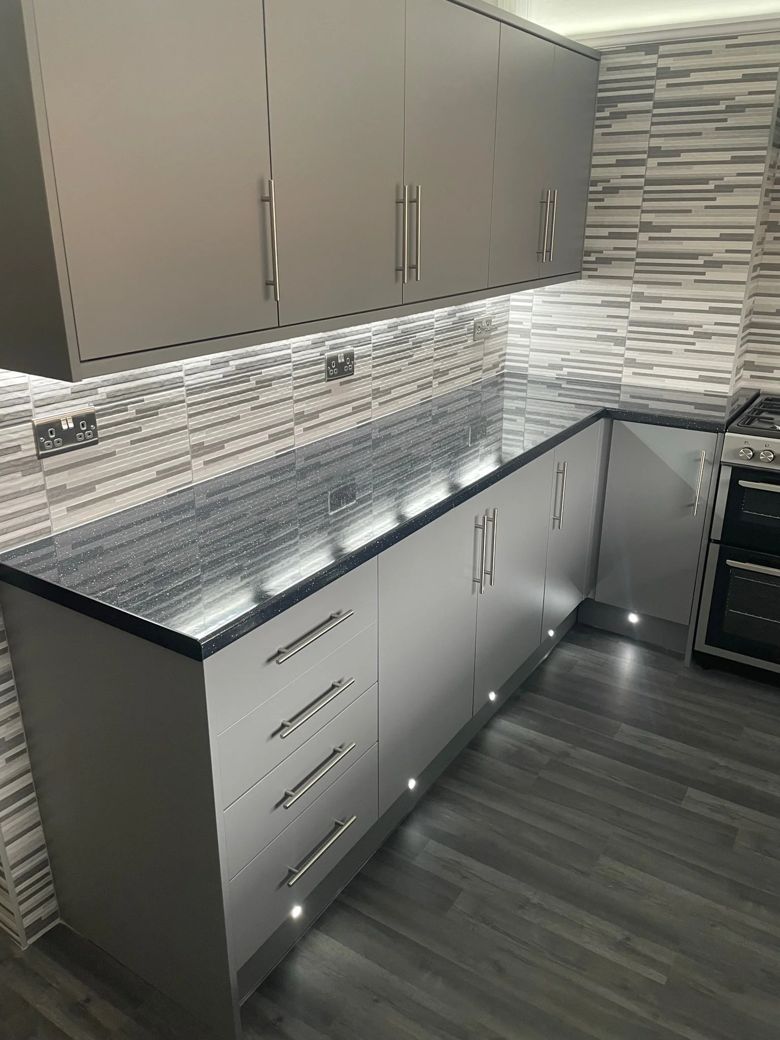 Modern kitchen with grey cabinets, a black speckled countertop, textured tile backsplash, and built-in lighting beneath the upper cabinets and along the bottom edge of the lower cabinets.