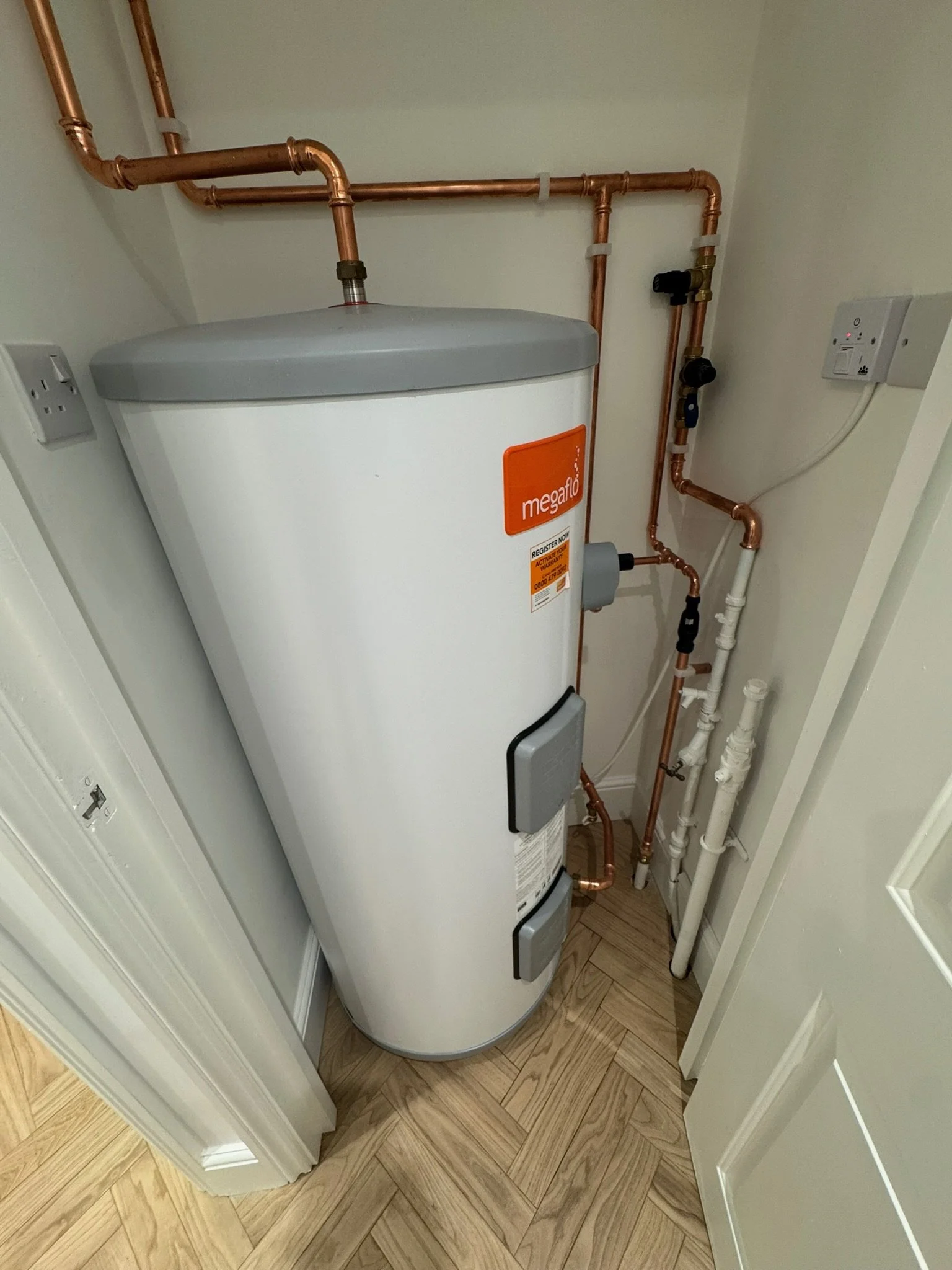 A white Megaflo hot water heater in a small closet with copper pipes and white PVC pipes connected to it.