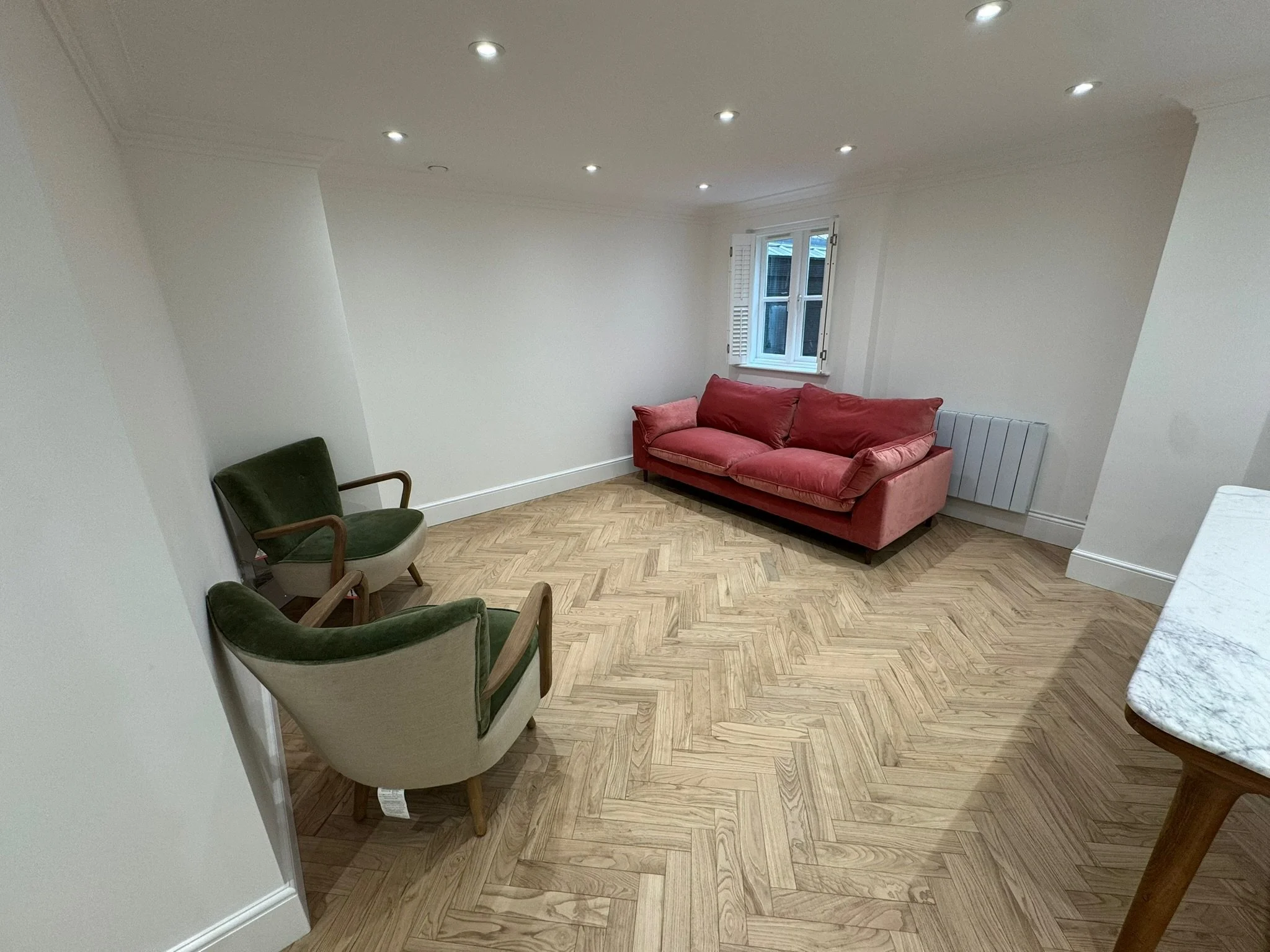 A living room with a hardwood herringbone floor, a red sofa, two old-fashioned armchairs with green and beige upholstery and wooden arms, white walls, a window with shutters, recessed ceiling lights, and a white radiator.