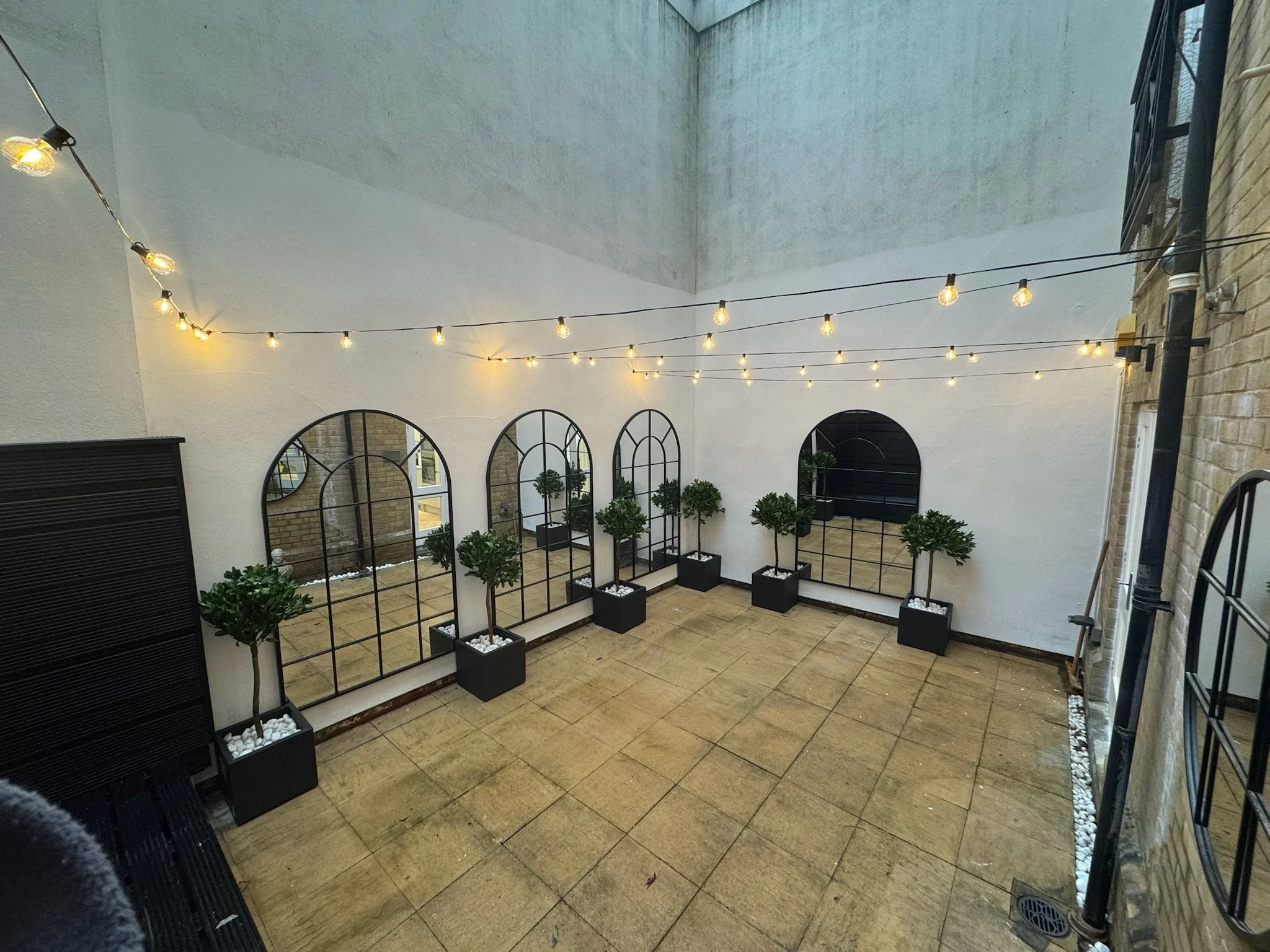 An outdoor patio with string lights, potted plants with trees, large mirrors, and a tiled floor.