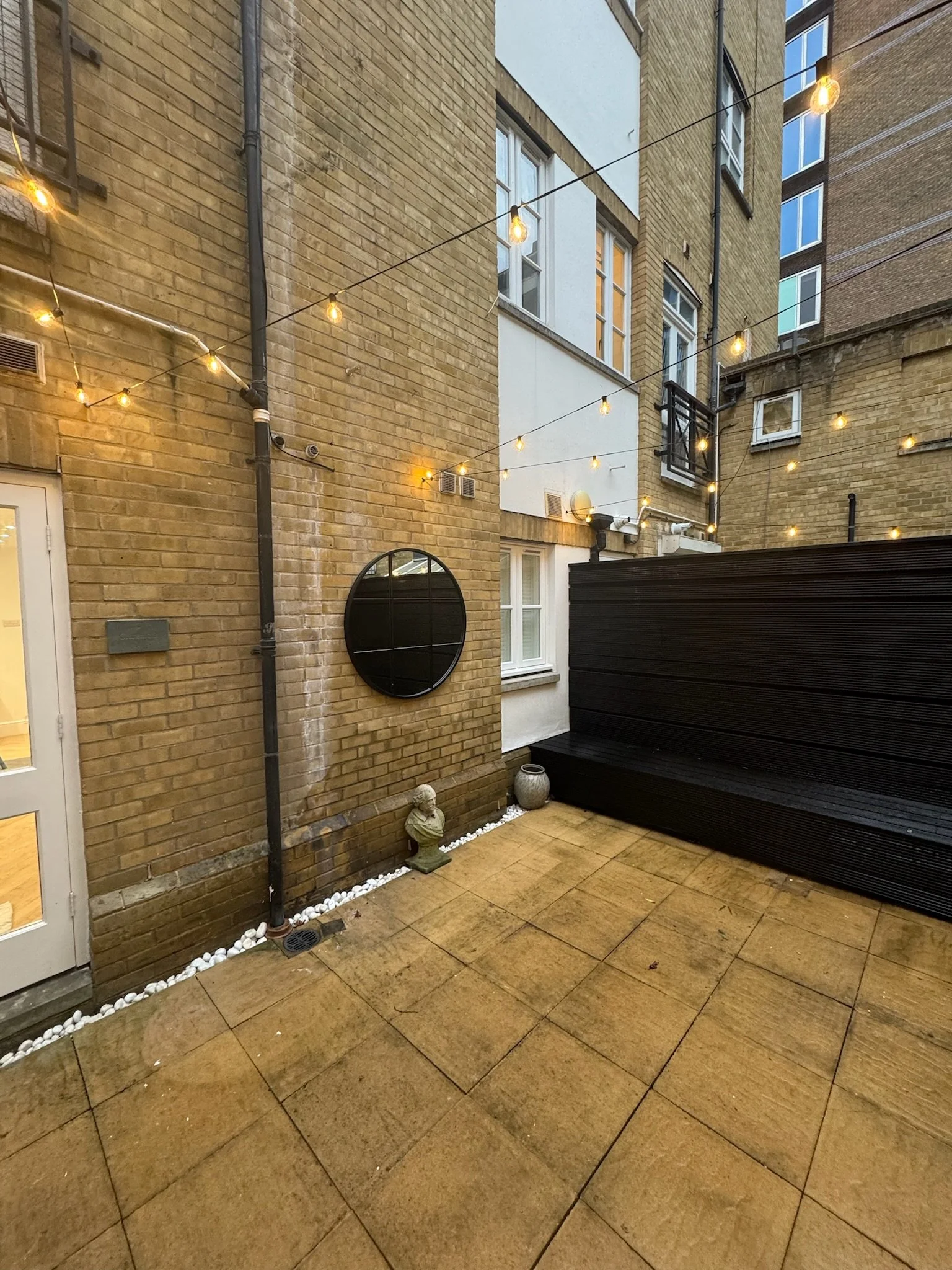 A small outdoor patio area with yellow brick walls, string lights overhead, and a black wooden privacy wall. There are a few decorative statues and planters, with a round mirror hanging on the wall.