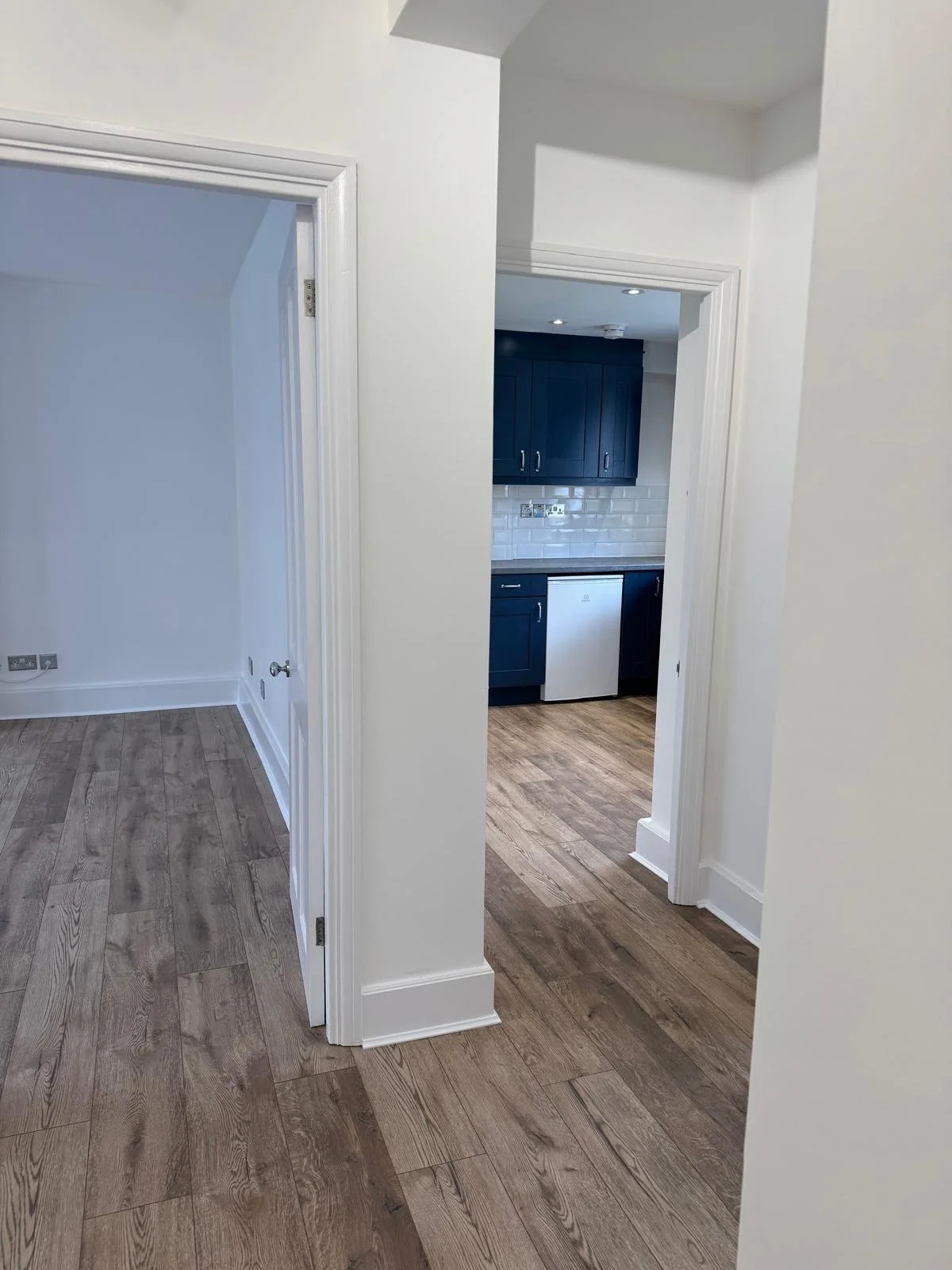 View of an interior space showing a hallway with white walls and wood flooring, leading to a kitchen with dark blue cabinets, a white subway tile backsplash, and a small white refrigerator.