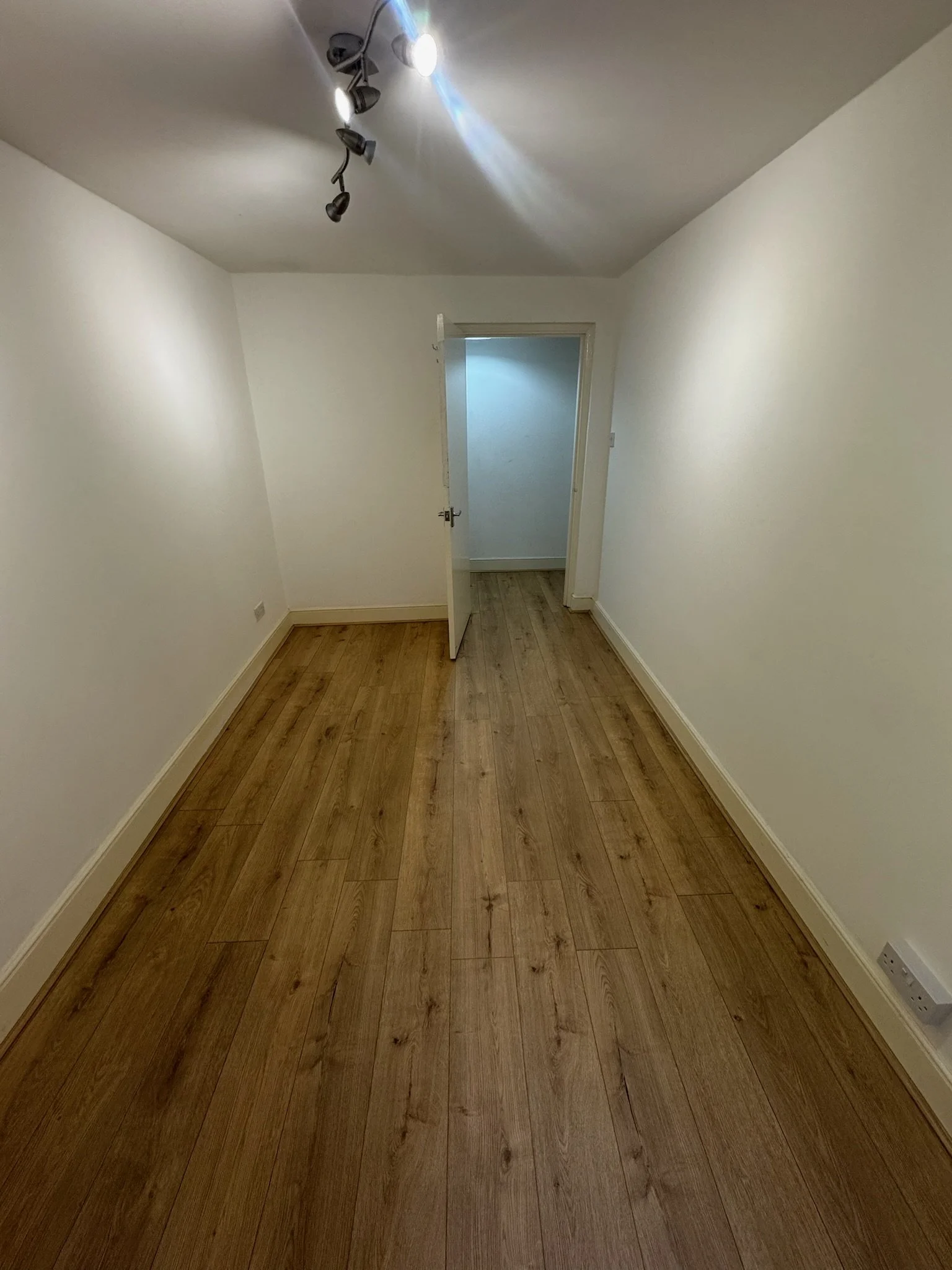An empty room with white walls, wooden flooring, a ceiling with a light fixture, and an open door leading to another room.