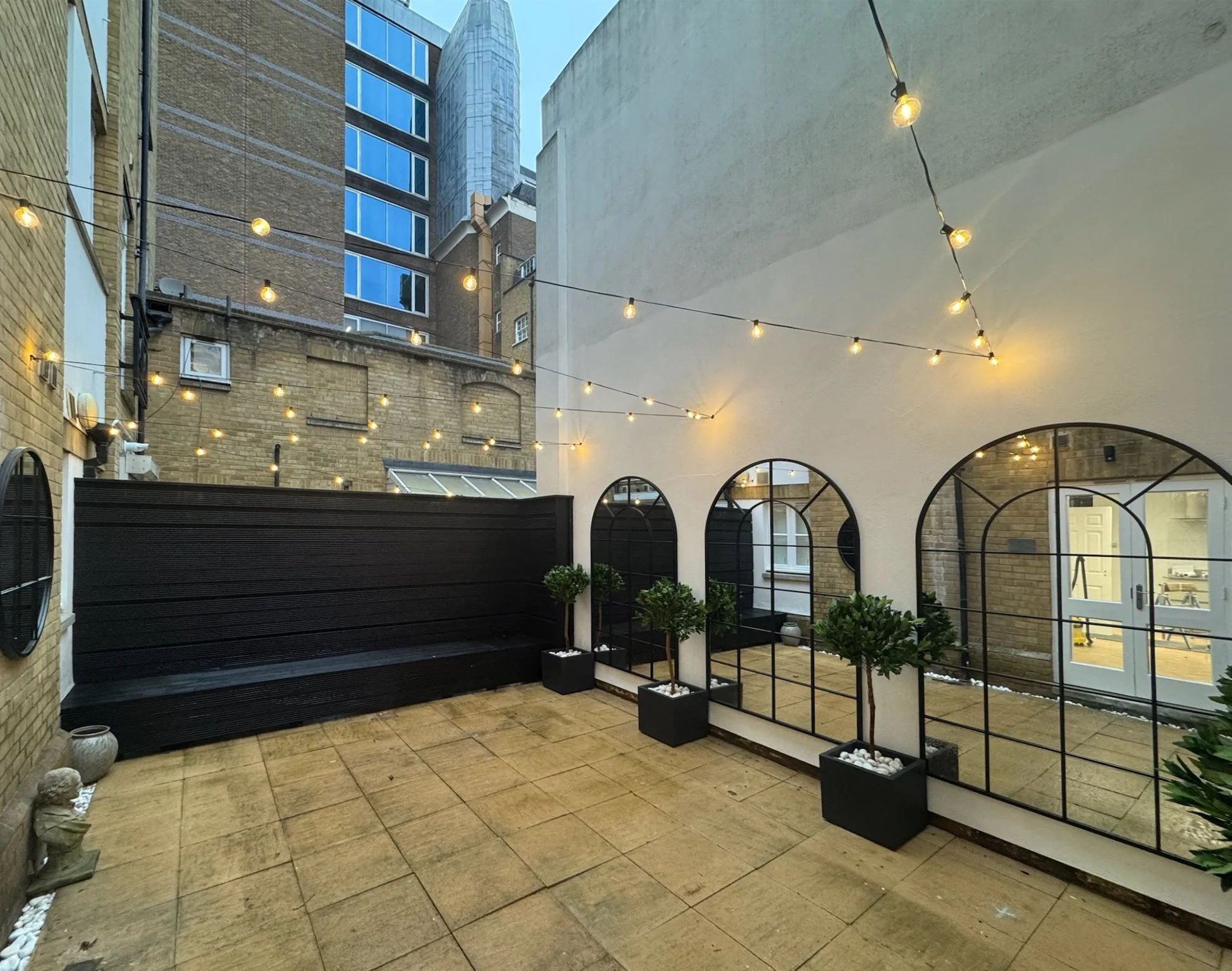 Urban outdoor patio with string lights, potted trees, and a black fence and lounge seating area, surrounded by tall buildings.
