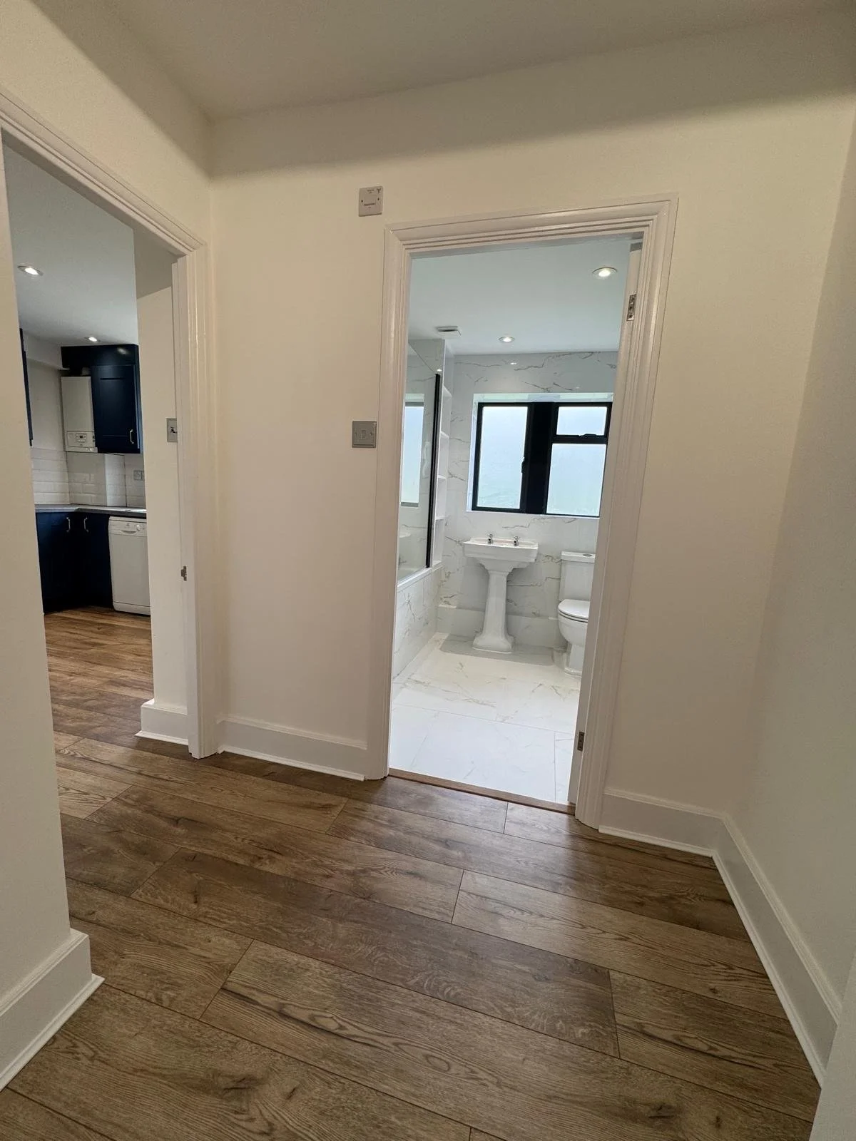 View of a hallway leading to a bathroom with a pedestal sink, toilet, and window, with a part of kitchen visible on the left, featuring wooden flooring and white walls.