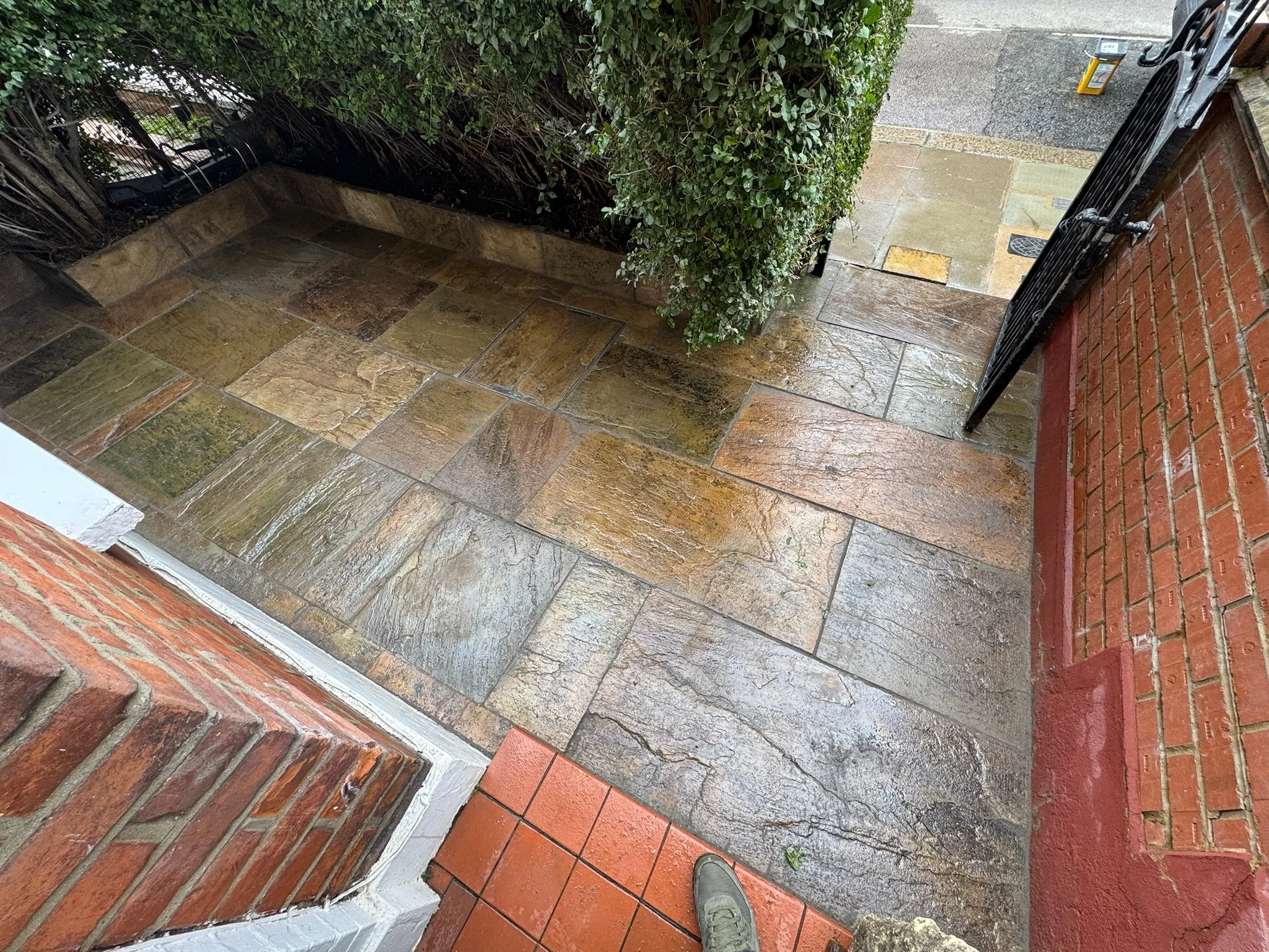 Wet stone patio on a porch with a brick step, a black metal railing, a green bush, and a wet sidewalk outside.