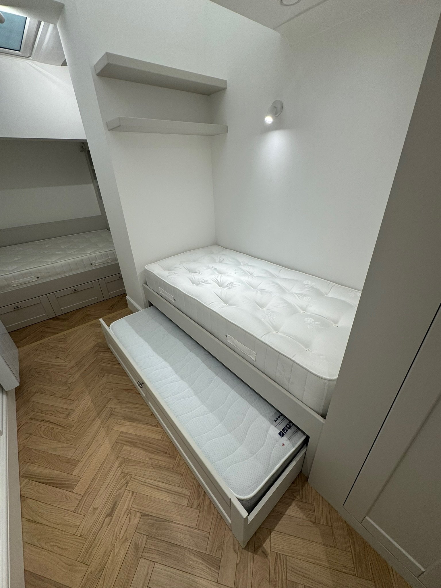 A small bedroom with a white trundle bed and mattress with wooden flooring and white walls. Two white shelves are mounted on the wall above the bed, and a small wall light is turned on.