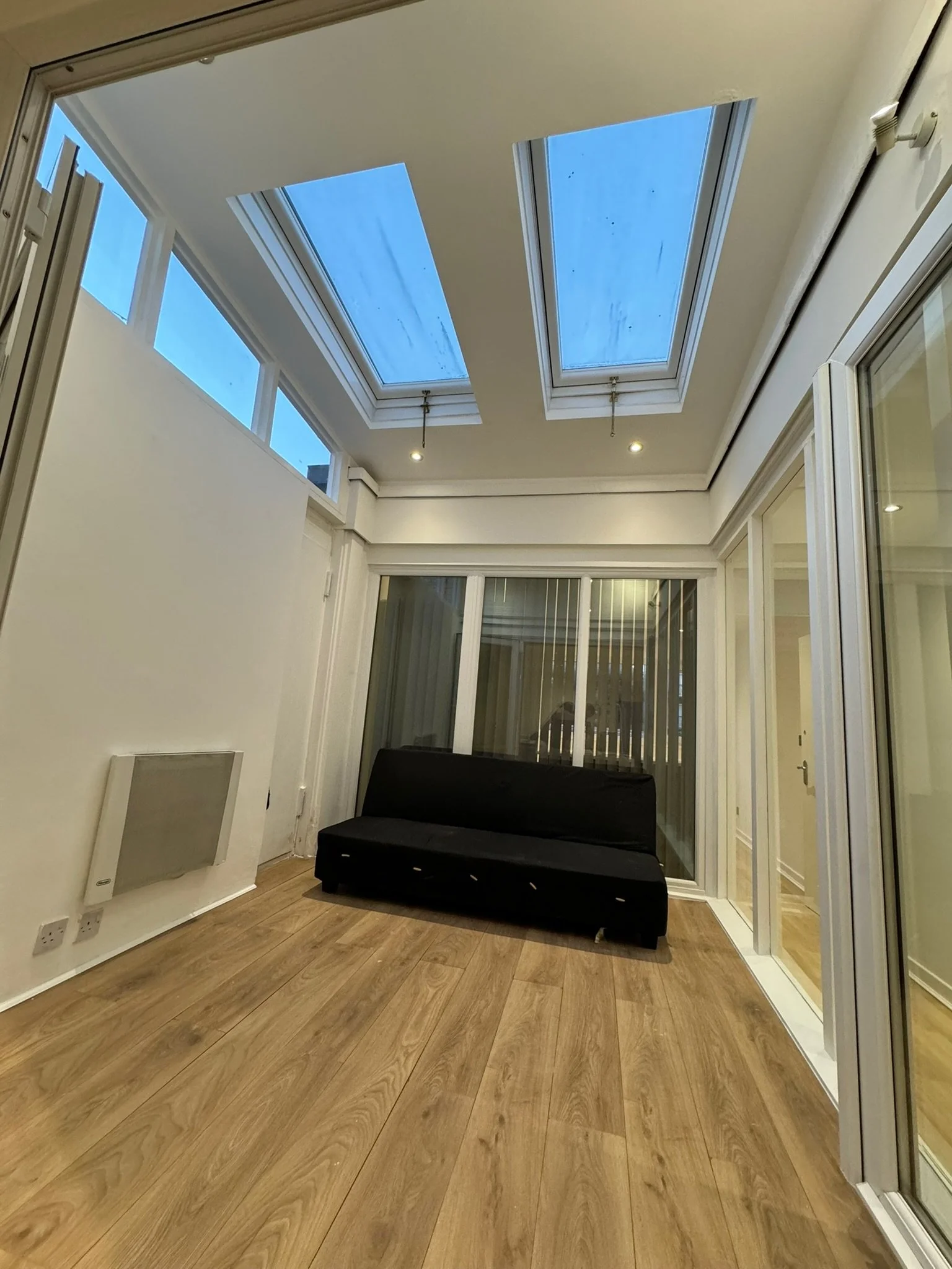 A sunroom with large skylights, wooden flooring, and glass sliding doors. There is a black couch against the back wall and some vertical blinds on the right. The ceiling has recessed lighting.