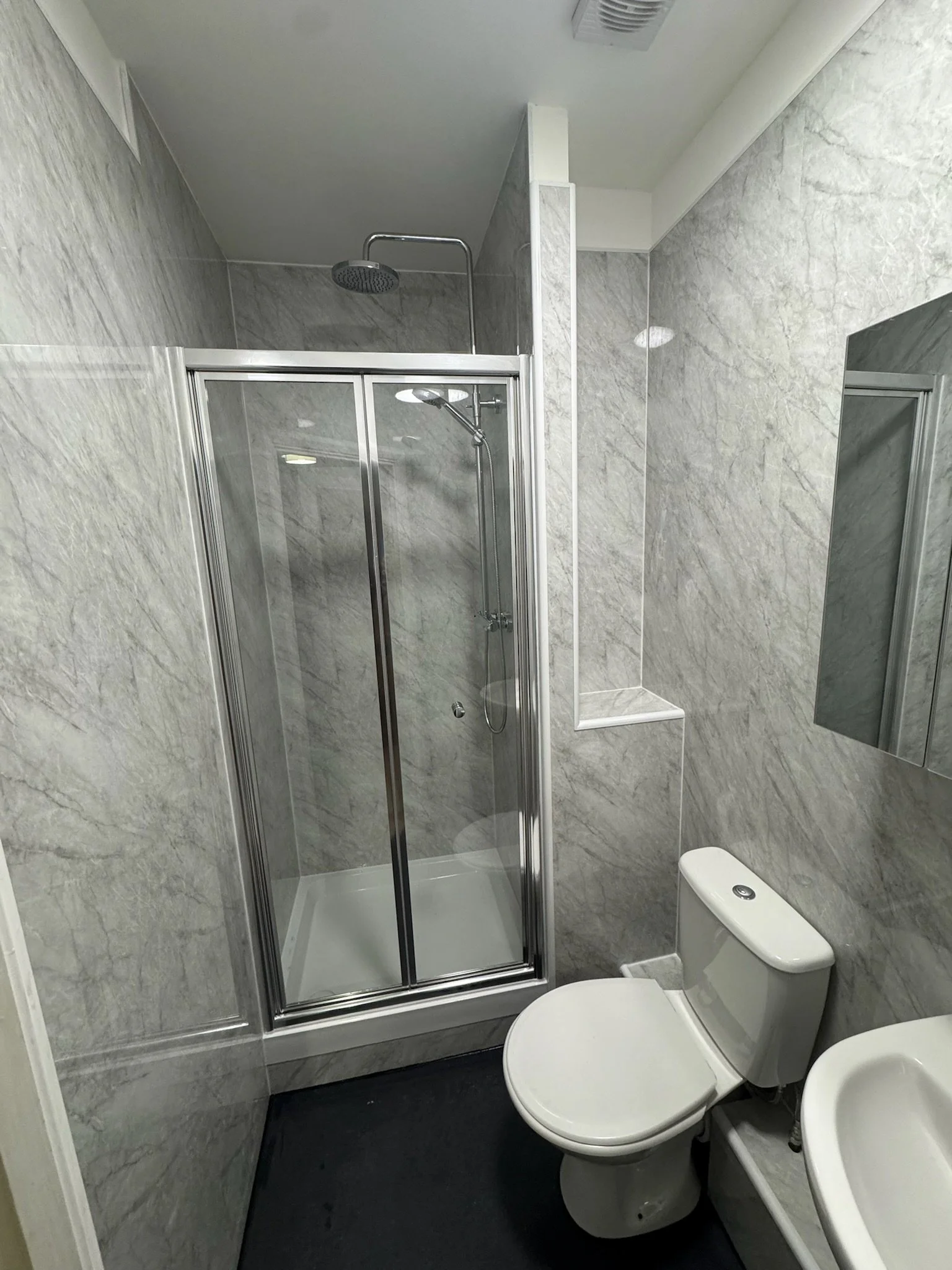 Small bathroom with a gray marble shower, a toilet, and a small sink, with gray marble walls and a black floor.