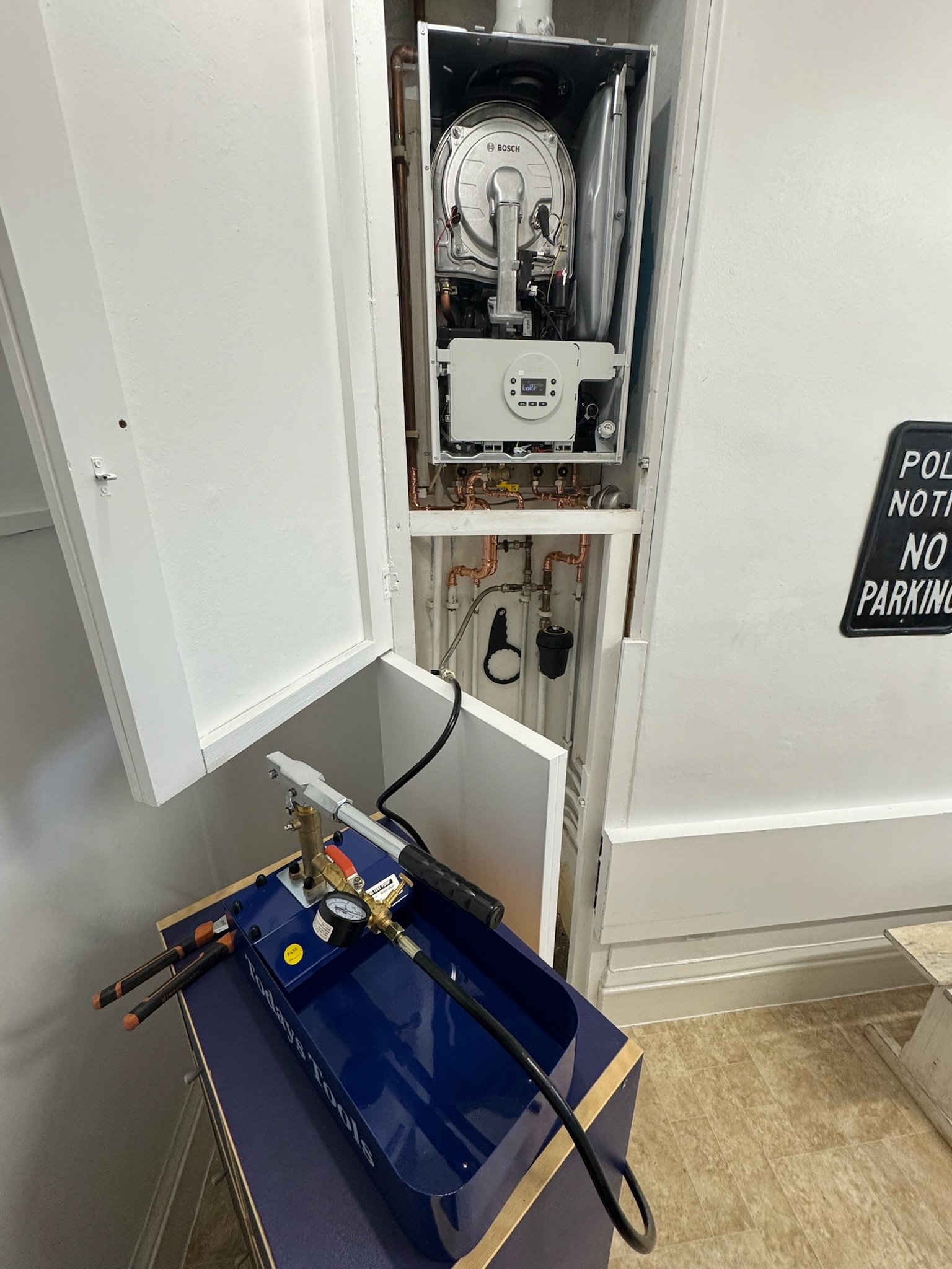 A technician's service tool with a black hose, gauge, and connectors on a blue tray is positioned in front of a boiler with an open access panel, revealing internal components and copper piping.