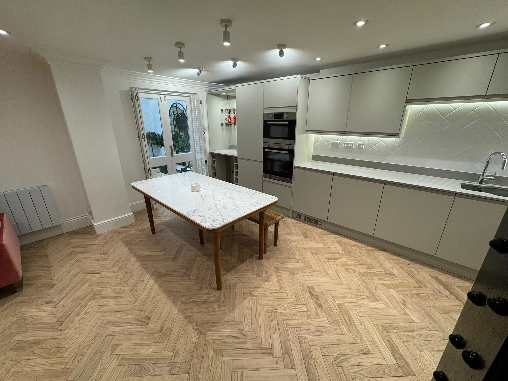 Modern kitchen with white cabinetry, built-in oven, marble-topped dining table, and hardwood floor.