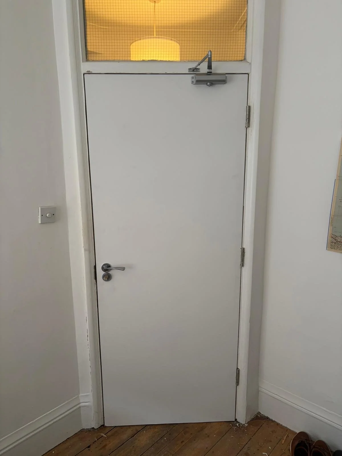 A plain white door with a silver handle and a lock, featuring a metal door closer at the top, and a small window with a wire mesh screen at the top allowing light to pass through.