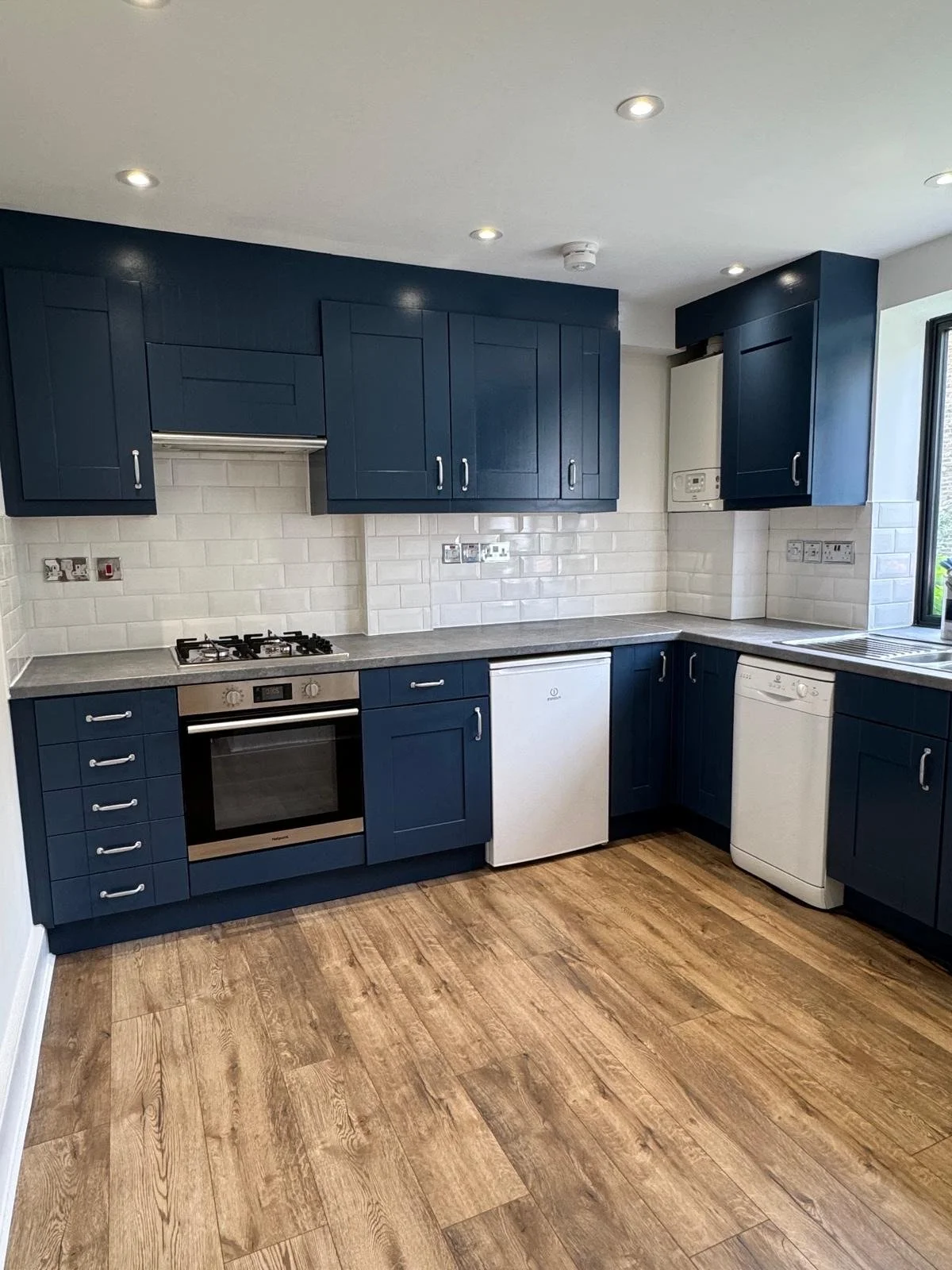 Modern kitchen with blue cabinets, white subway tile backsplash, white appliances, and wood flooring.