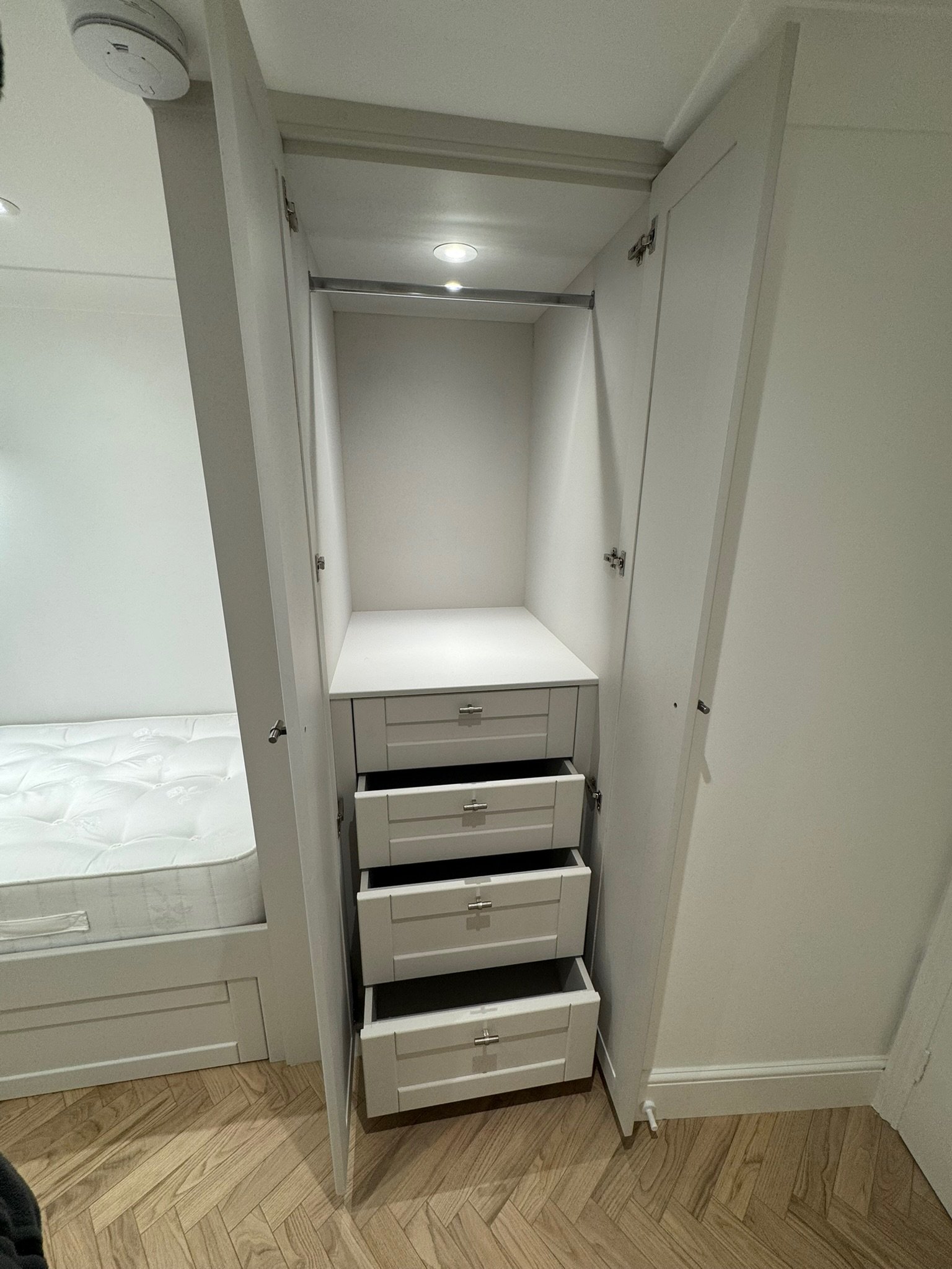 Empty white wardrobe with three pull-out drawers and closed side cabinets, located next to a bed with a white mattress, in a room with wooden flooring.