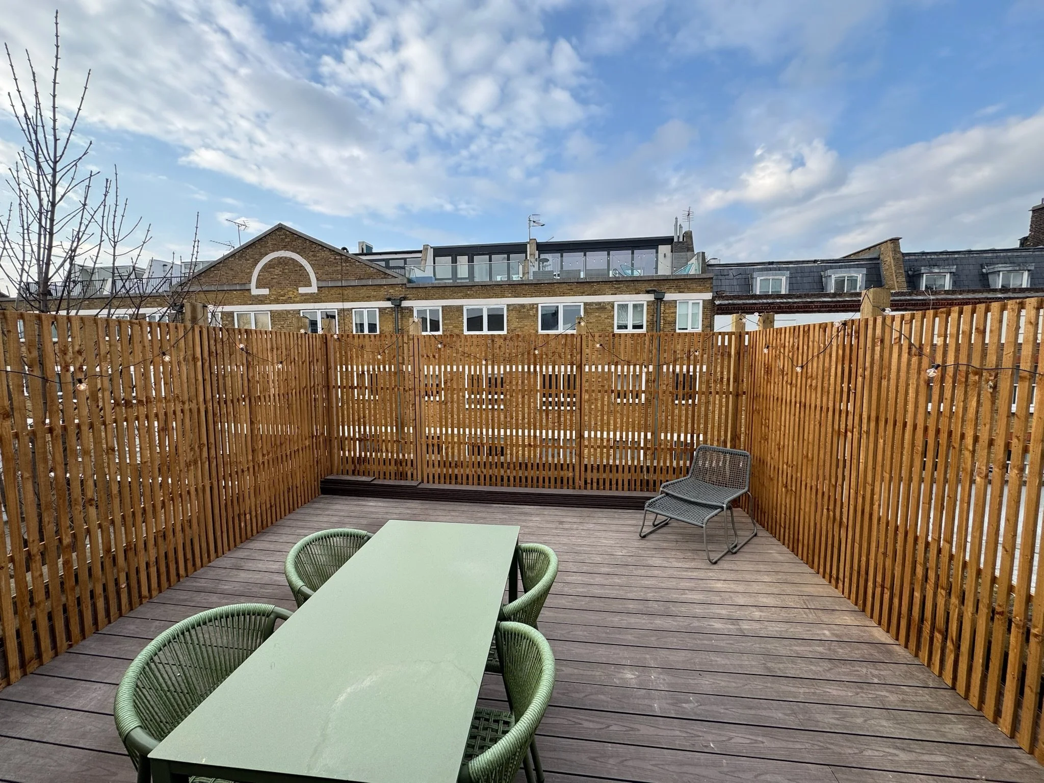 Empty rooftop patio with wooden deck, green table, four green chairs, a gray chair, and a wooden fence, with multi-story buildings and a partly cloudy sky in the background.