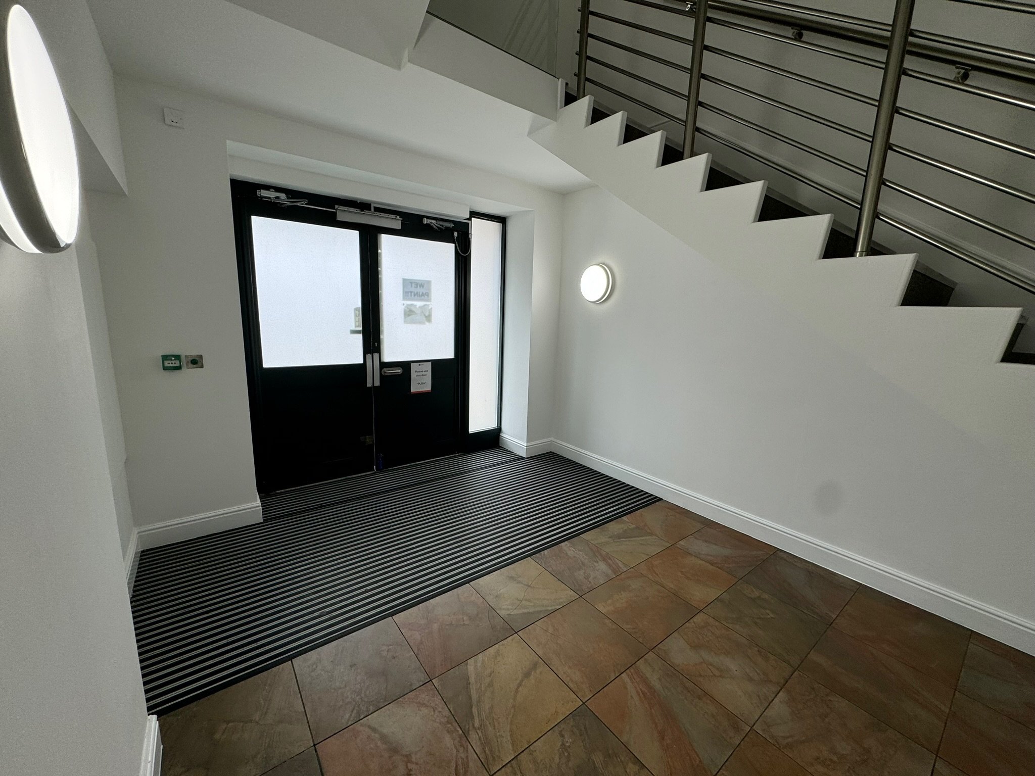 Interior view of a building lobby with double glass doors, a striped rubber doormat, tiled floor, white walls, and a metal staircase with railings.