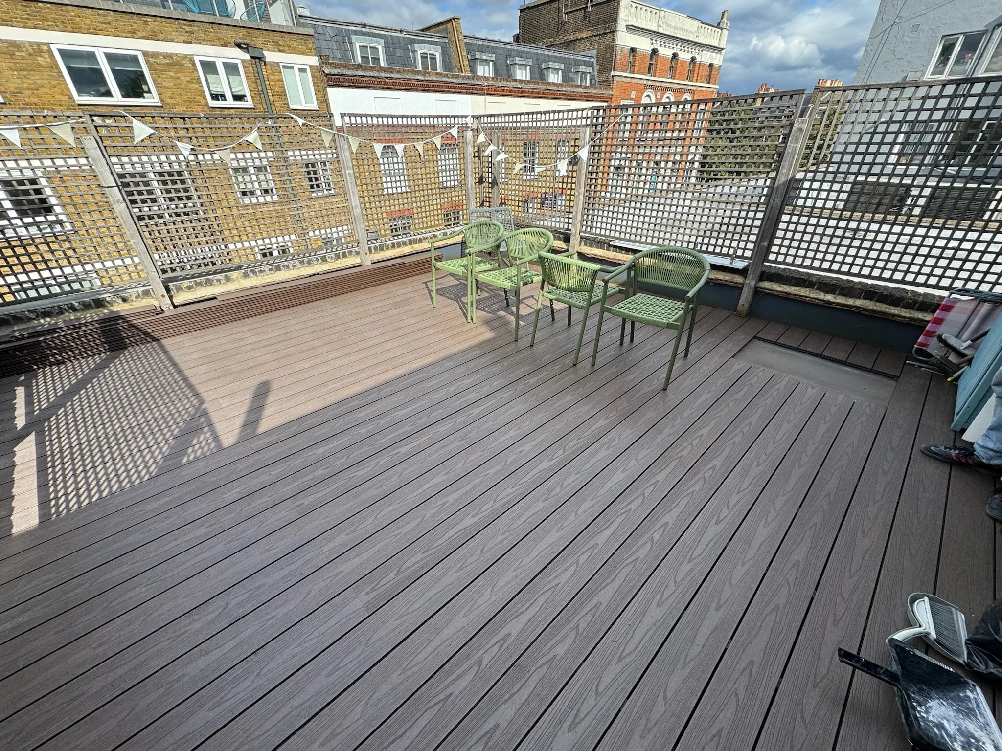 An outdoor rooftop terrace with wooden decking, surrounded by a lattice fence. Four green chairs are arranged in the corner, with one slightly different in style. In the background, multi-story brick and concrete buildings are visible under a partly 