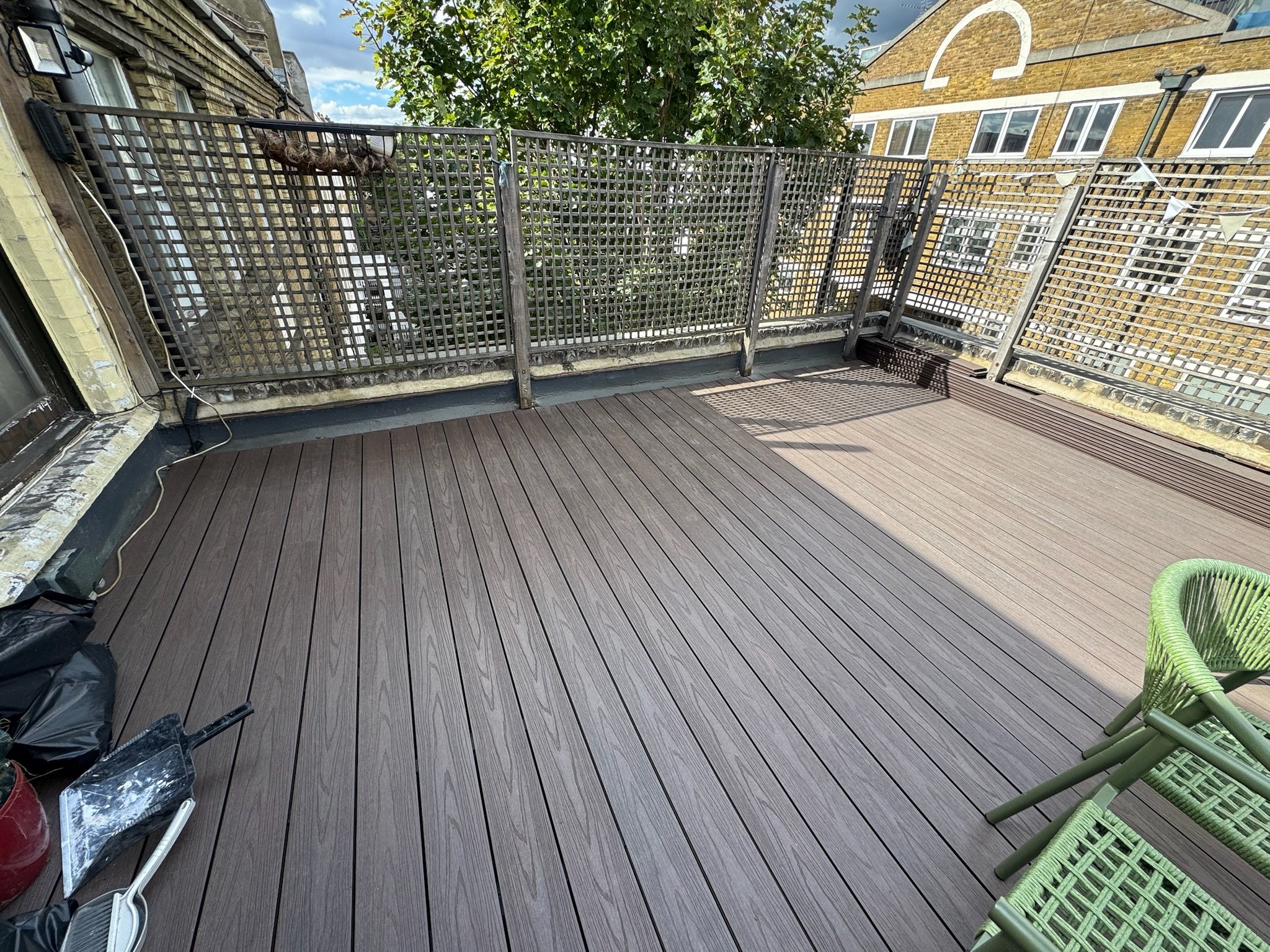 A rooftop balcony with brown wooden decking, green chairs, and a metal lattice railing. There are some tools and tools bags in the corner. The sky is partly cloudy and there's a tree visible over the railing.