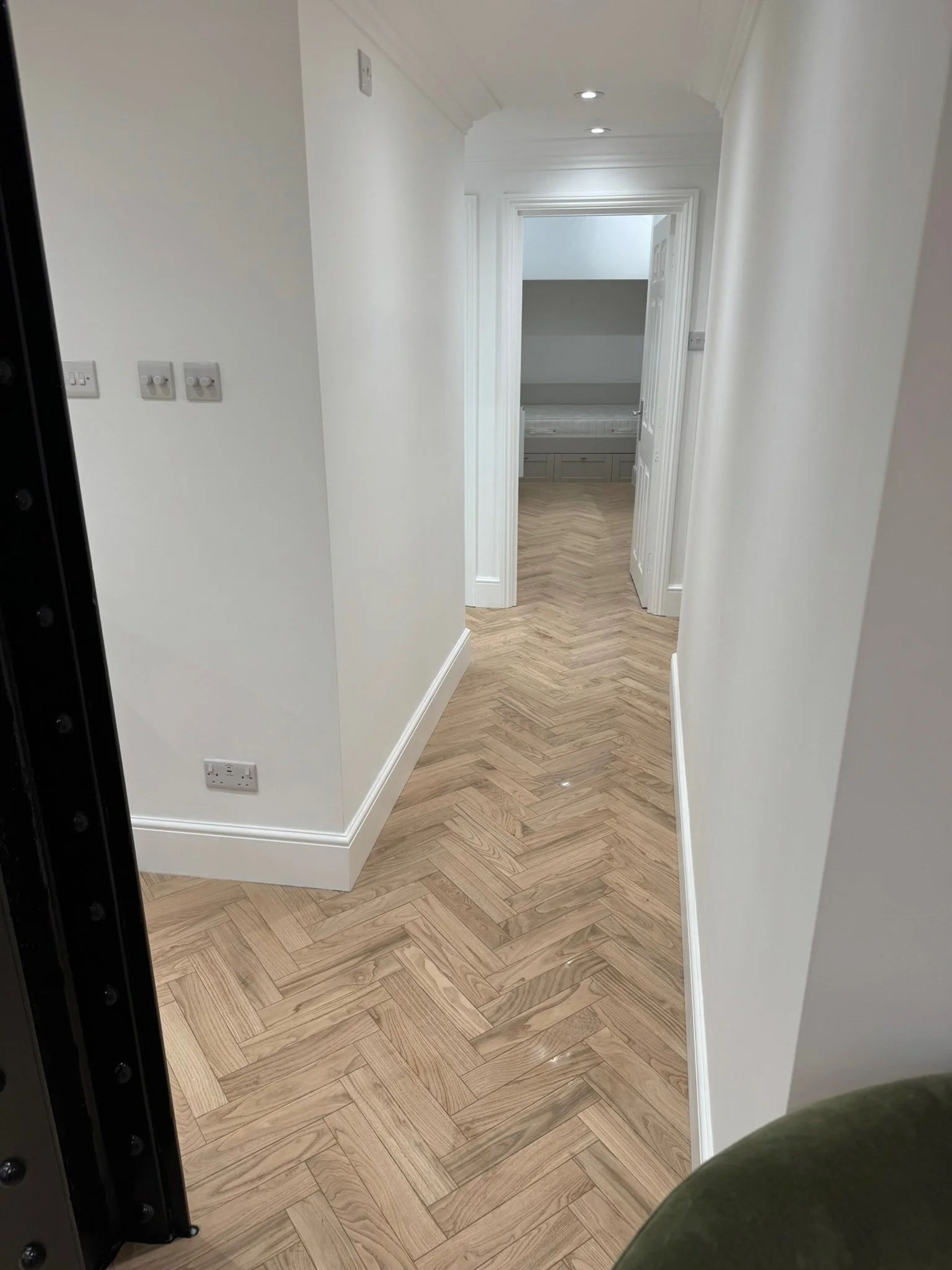 An interior hallway with light-colored walls, white crown molding, and a herringbone-patterned wooden floor leading to a slightly open door at the end.