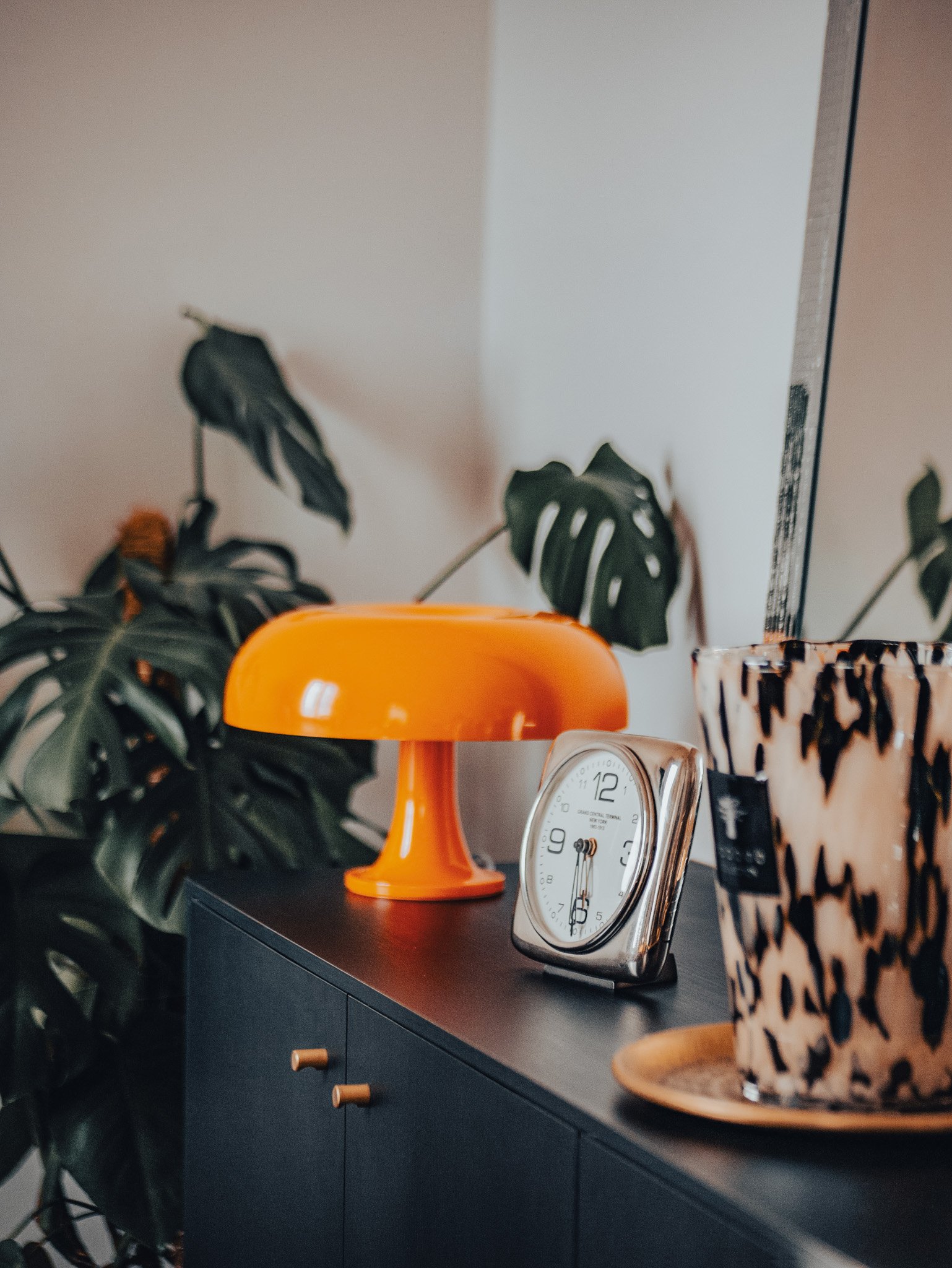 Une table noire avec une plante verte en arrière-plan, une lampe orange, une horloge à chiffres romains et un pot décoratif à motifs noirs et beige sur un plateau en bois.
