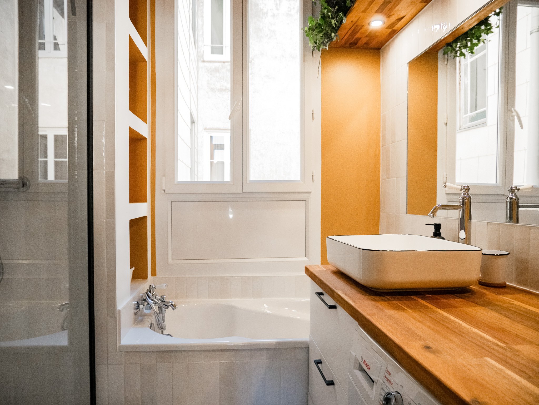 Rénovation d'un salle de bain dans un appartement ancien à Rouen. Meubles sur-mesure avec étagères et placards en bois foncé, douche à l'italienne et baignoire avec huisseries noires, peinture couleur cumin, miroir sur mesure avec zellige mural blanc