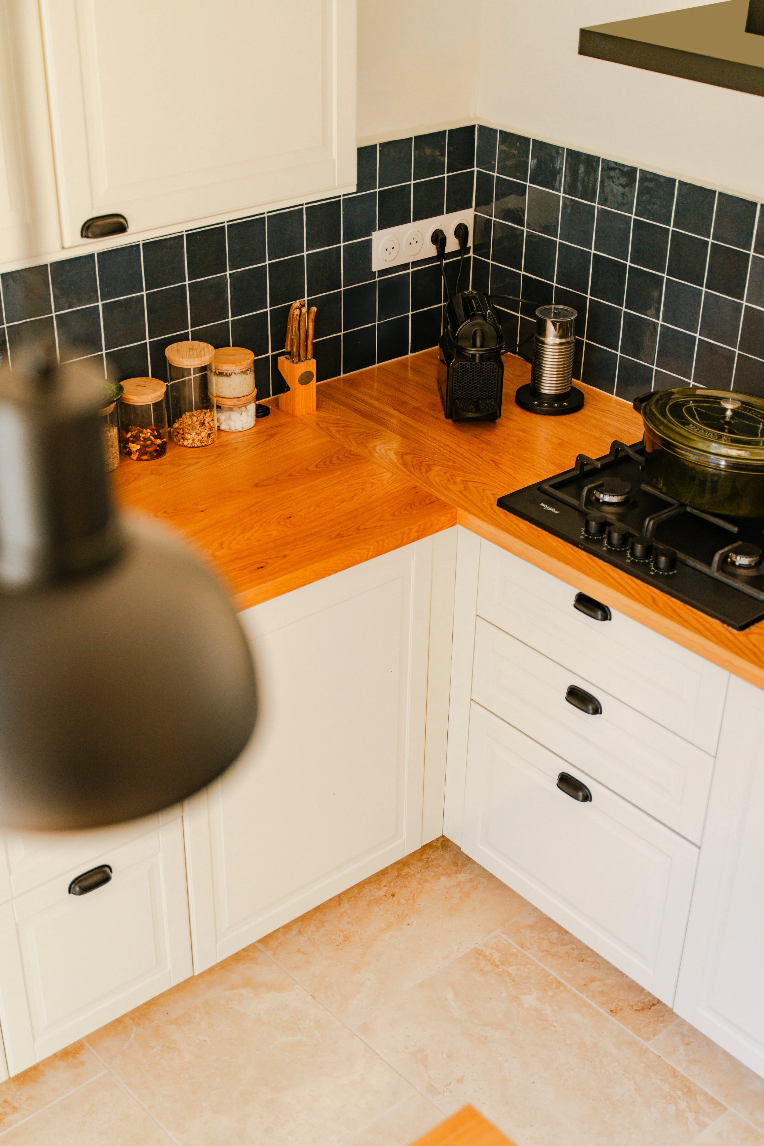 Vue partielle d'une cuisine avec plan de travail en bois clair, prises électriques, ustensiles, un café avec machine, et un feu de cuisine, avec sol en carrelage beige
