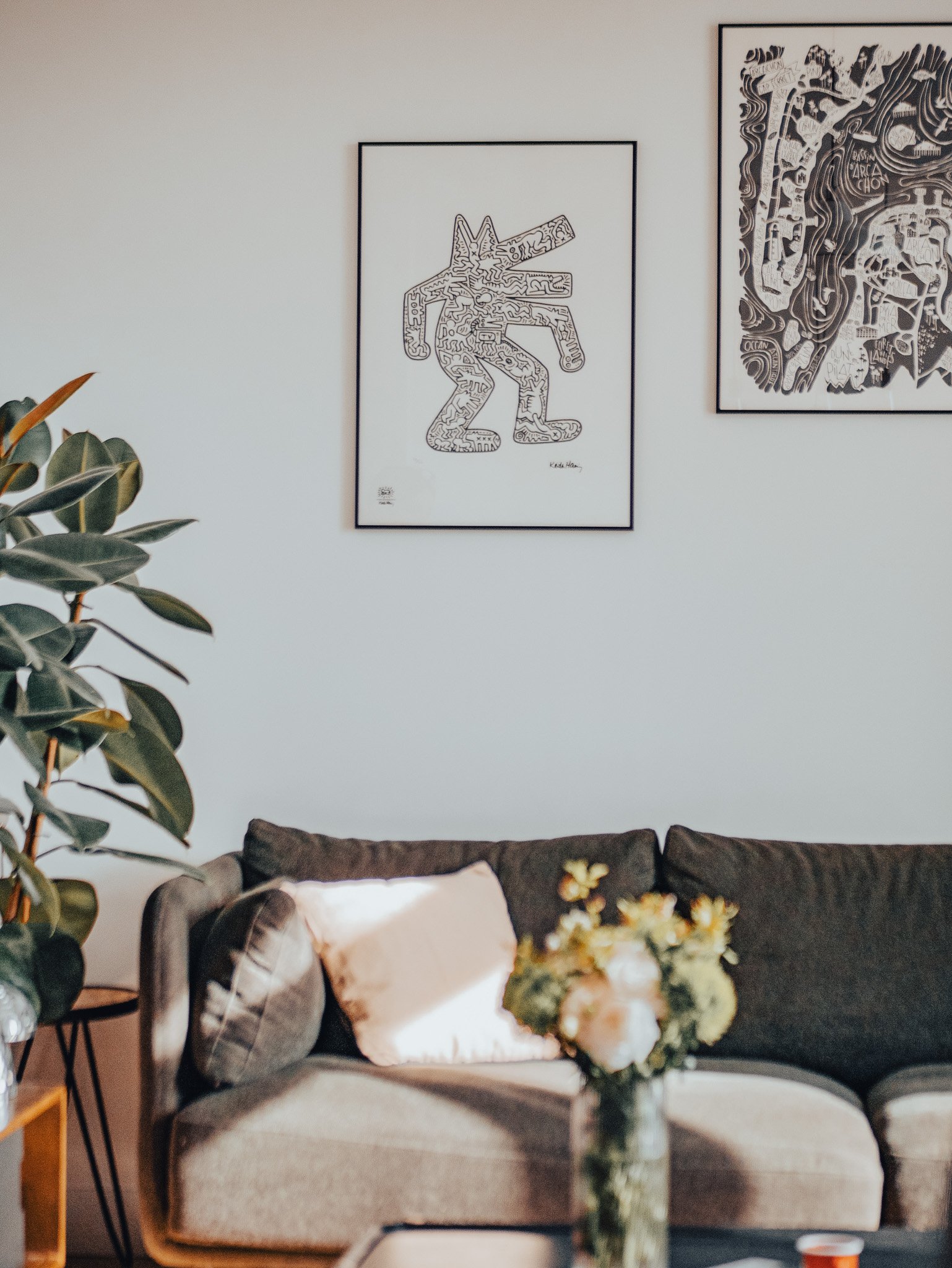 Salon avec un canapé gris, un coussin blanc, une plante à gauche, une table avec un vase de fleurs, et deux œuvres d'art encadrées en noir et blanc sur le mur.